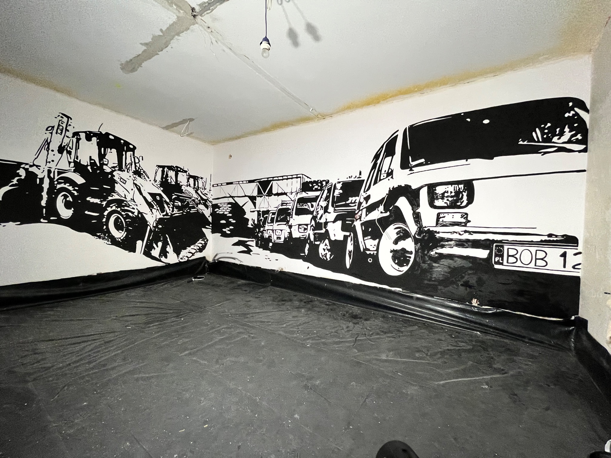 Czarno-białe malowidło na ścianie przedstawiające ciągniki i samochody, z folią ochronną na podłodze w trakcie remontu.