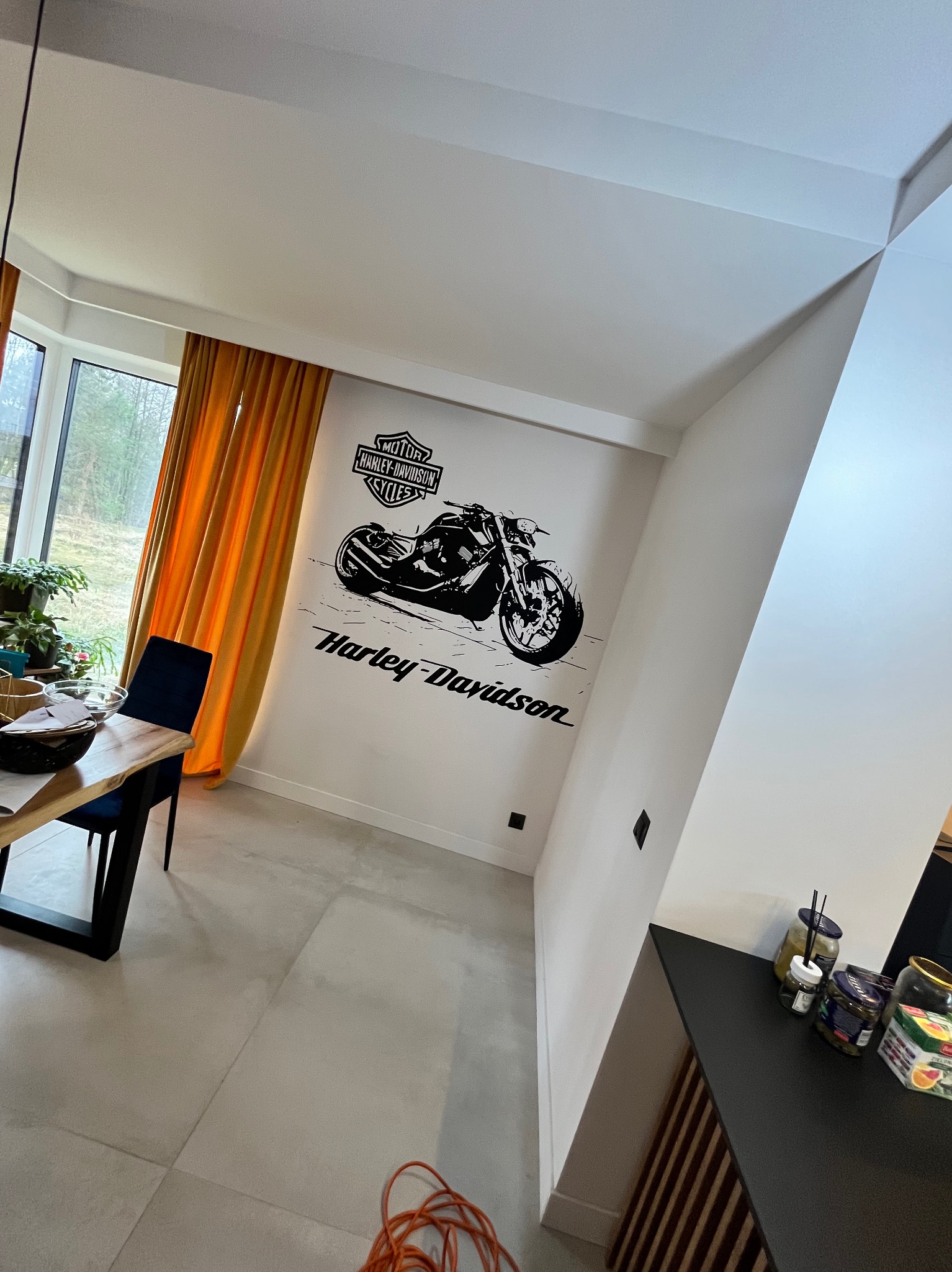 Biała ściana z czarnym motywem motocykla Harley-Davidson w nowoczesnym salonie z pomarańczowymi zasłonami i minimalistycznym wystrojem.