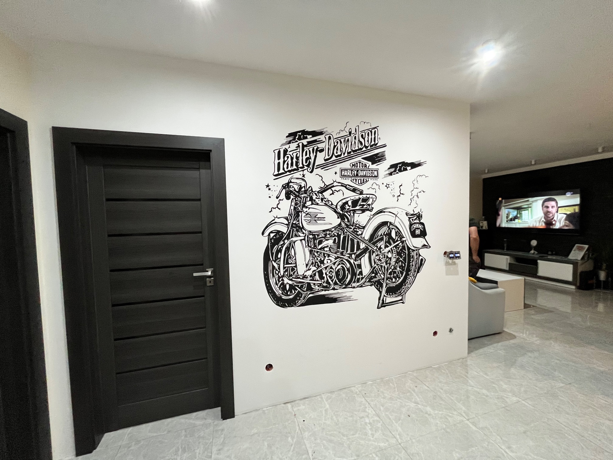Ściana w salonie z czarnym rysunkiem motocykla Harley-Davidson obok ciemnych drzwi, widoczne niedokończone gniazdka elektryczne i szare płytki podłogowe.