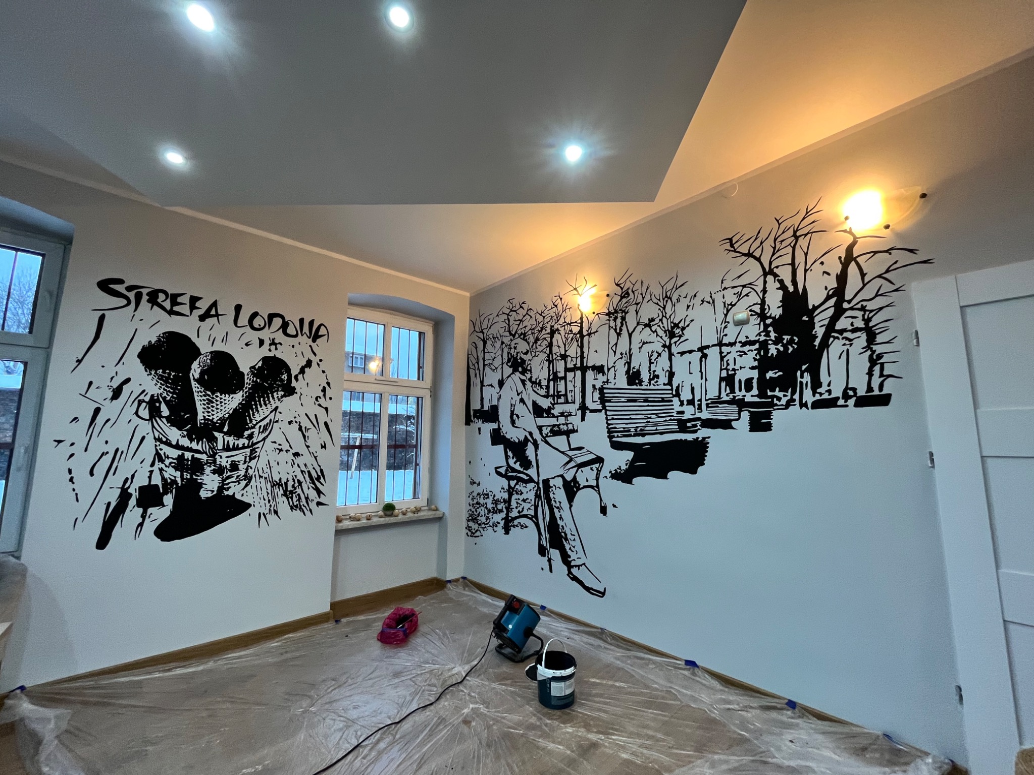Pokój z czarno-białymi malowidłami na ścianach: napis 'Strefa Lodowa' z grafiką lodów oraz scena parkowa z osobą siedzącą na ławce, widoczne narzędzia malarskie i folia ochronna na podłodze.