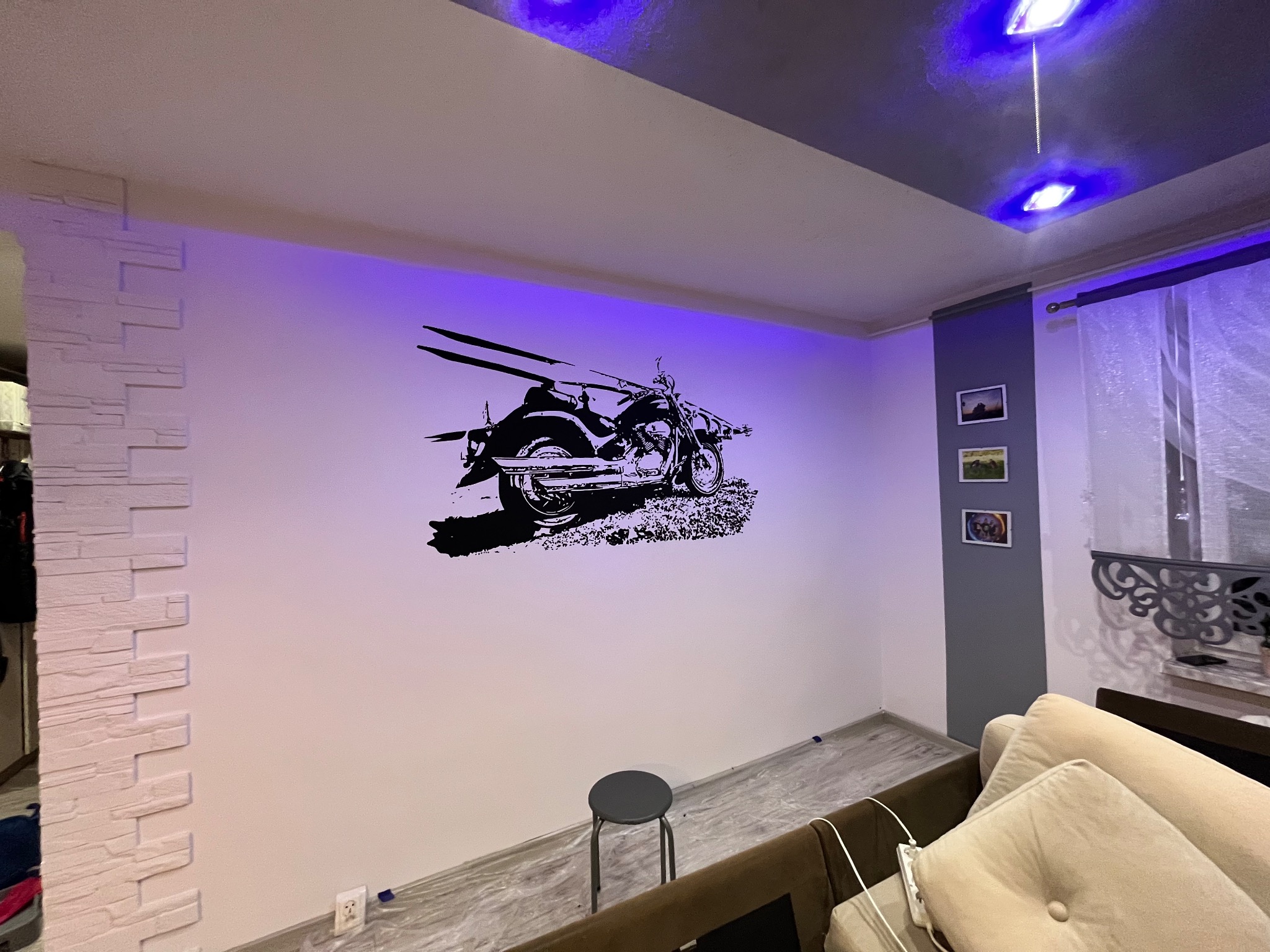 Ściana w pokoju z naklejoną czarną grafiką motocykla, oświetlona niebieskim światłem z sufitu, widoczny fragment dekoracyjnej ściany z imitacji kamienia oraz szary taboret.