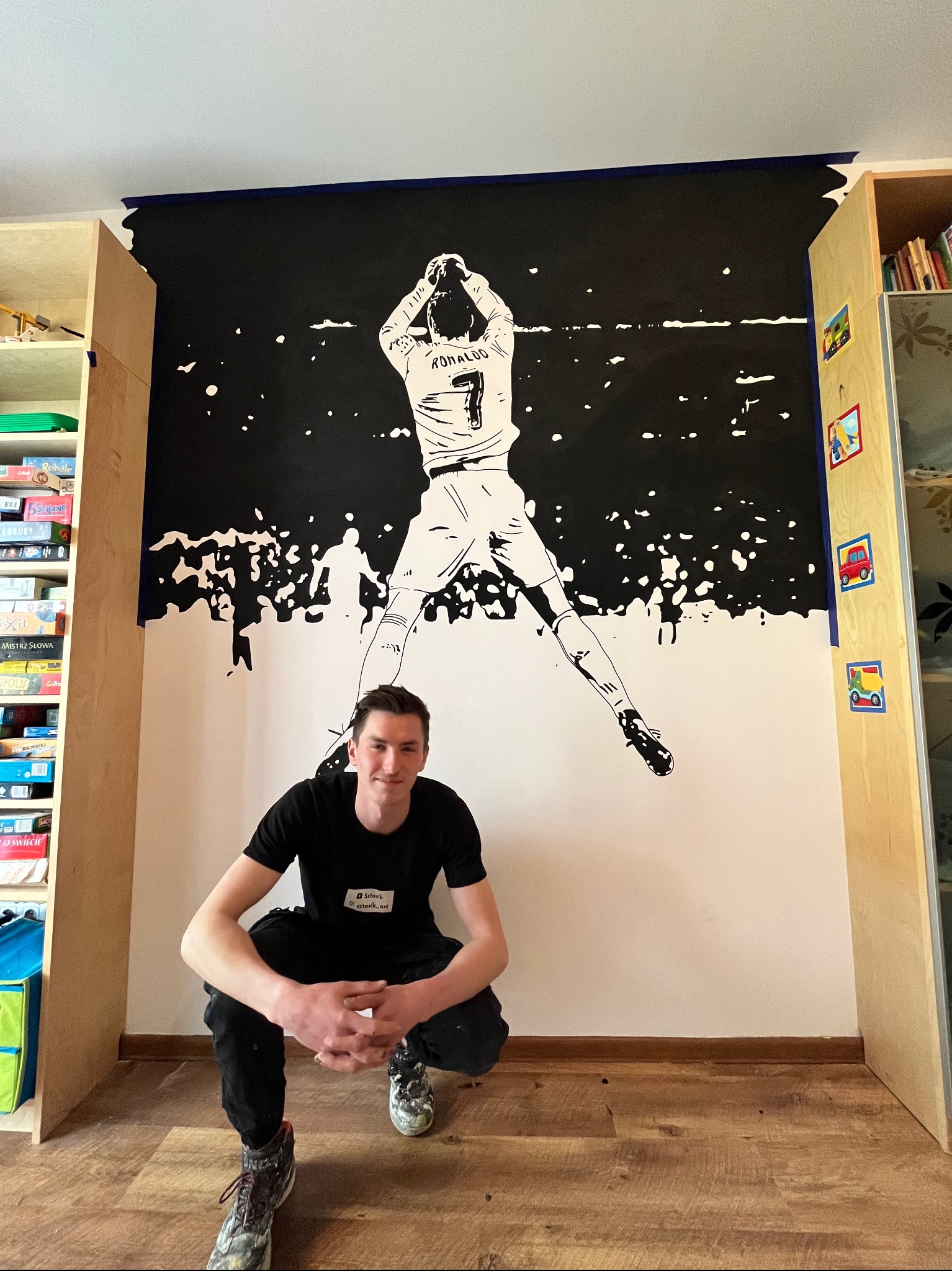 Młody mężczyzna kuca przed ścianą z ręcznie malowanym, czarno-białym muralem piłkarza Cristiano Ronaldo, praca w trakcie realizacji z widoczną taśmą malarską.