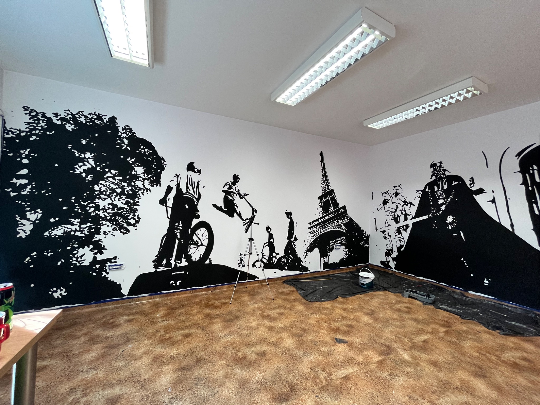 Czarno-białe malowidło ścienne przedstawiające rowerzystów, drzewo, wieżę Eiffla i postać Dartha Vadera, wykonane w biurze, z zabezpieczoną podłogą i widocznymi lampami sufitowymi.
