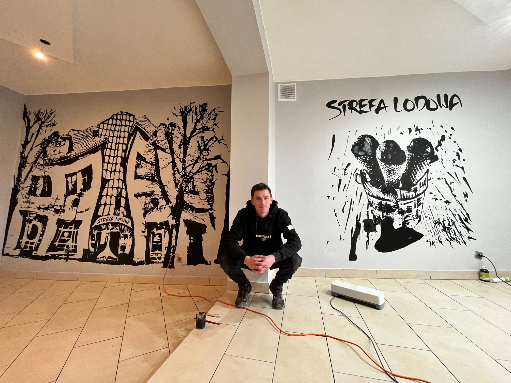 Artysta siedzi na kafelkach, prezentując ukończone malowidła ścienne w stylu czarno-białym z motywem lodziarni 'Strefa Lodowa', widoczne pędzle i kabel zasilający