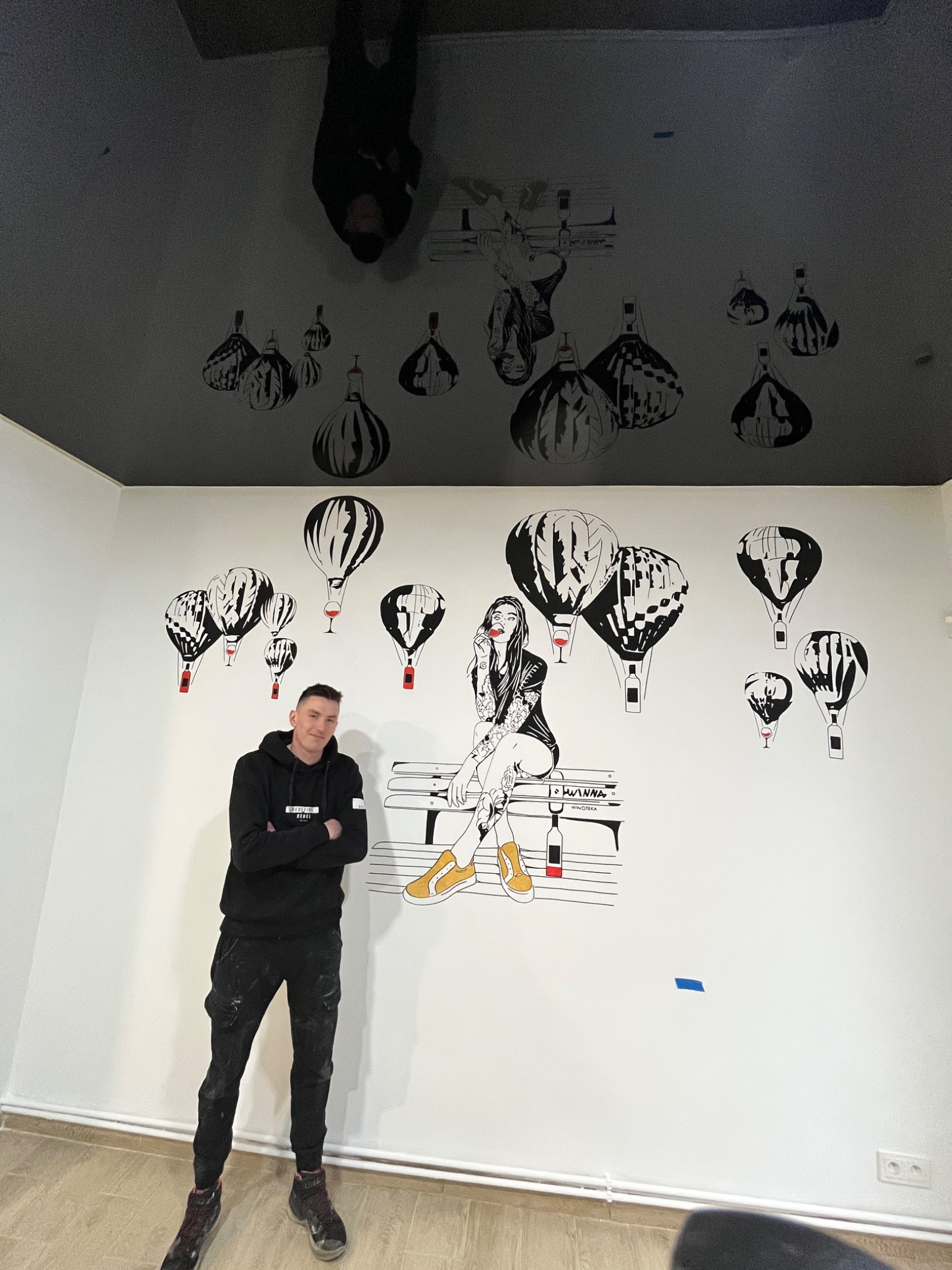 Mężczyzna w czarnej bluzie i spodniach stoi na tle ściany z czarno-białym rysunkiem balonu na gorące powietrze i kobiety siedzącej na ławce z butelką wina, wzór powtórzony na czarnym suficie.
