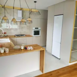 Jasna kuchnia na wymiar z drewnianym blatem i wyspą, oświetlona wiszącymi lampami z abażurami z rattanu, widok na zabudowę kuchenną z piekarnikiem i lodówką.