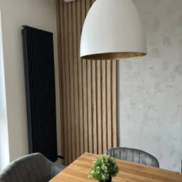 Drewniany blat stołu z małą rośliną doniczkową, szare krzesła tapicerowane, ściana z drewnianych paneli pionowych i dekoracyjna lampa wisząca w nowoczesnym wnętrzu.