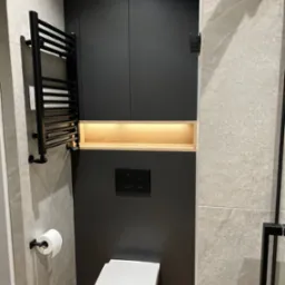 Nowoczesna łazienka z czarnymi szafkami w zabudowie, podświetlaną wnęką, grzejnikiem drabinkowym i wiszącą toaletą.