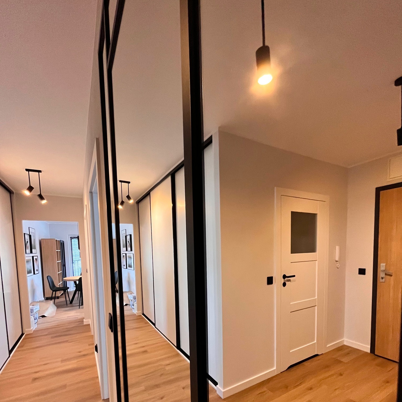 Korytarz po remoncie z jasną podłogą, białymi ścianami i czarnymi akcentami w postaci ram drzwi przesuwnych i oświetlenia, widok na salon z czarnymi krzesłami.
