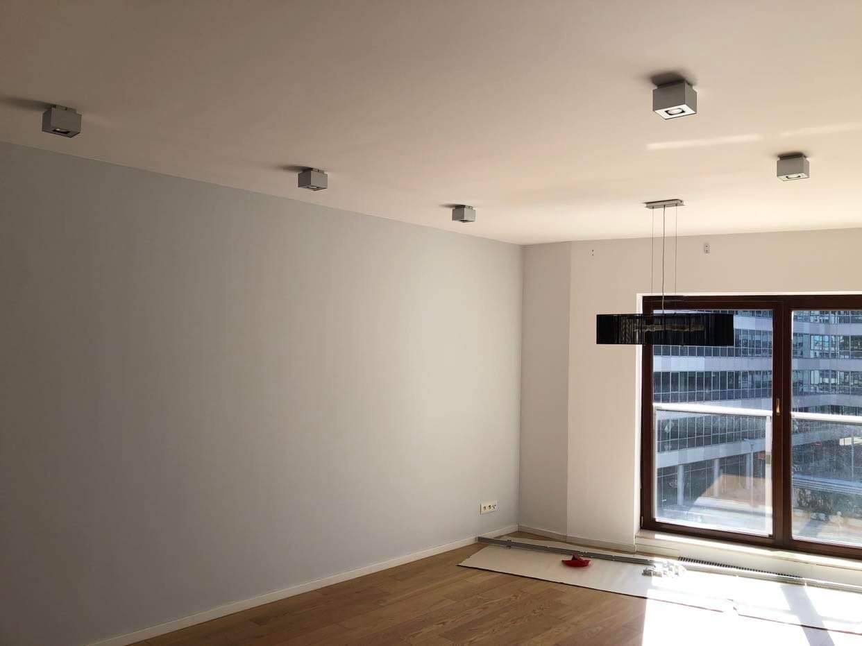 Jasne wnętrze pokoju po odświeżeniu ścian, z drewnianą podłogą, nowoczesnym oświetleniem sufitowym oraz dużym oknem z widokiem na budynek w oddali. Na podłodze widoczne zabezpieczenia przed...