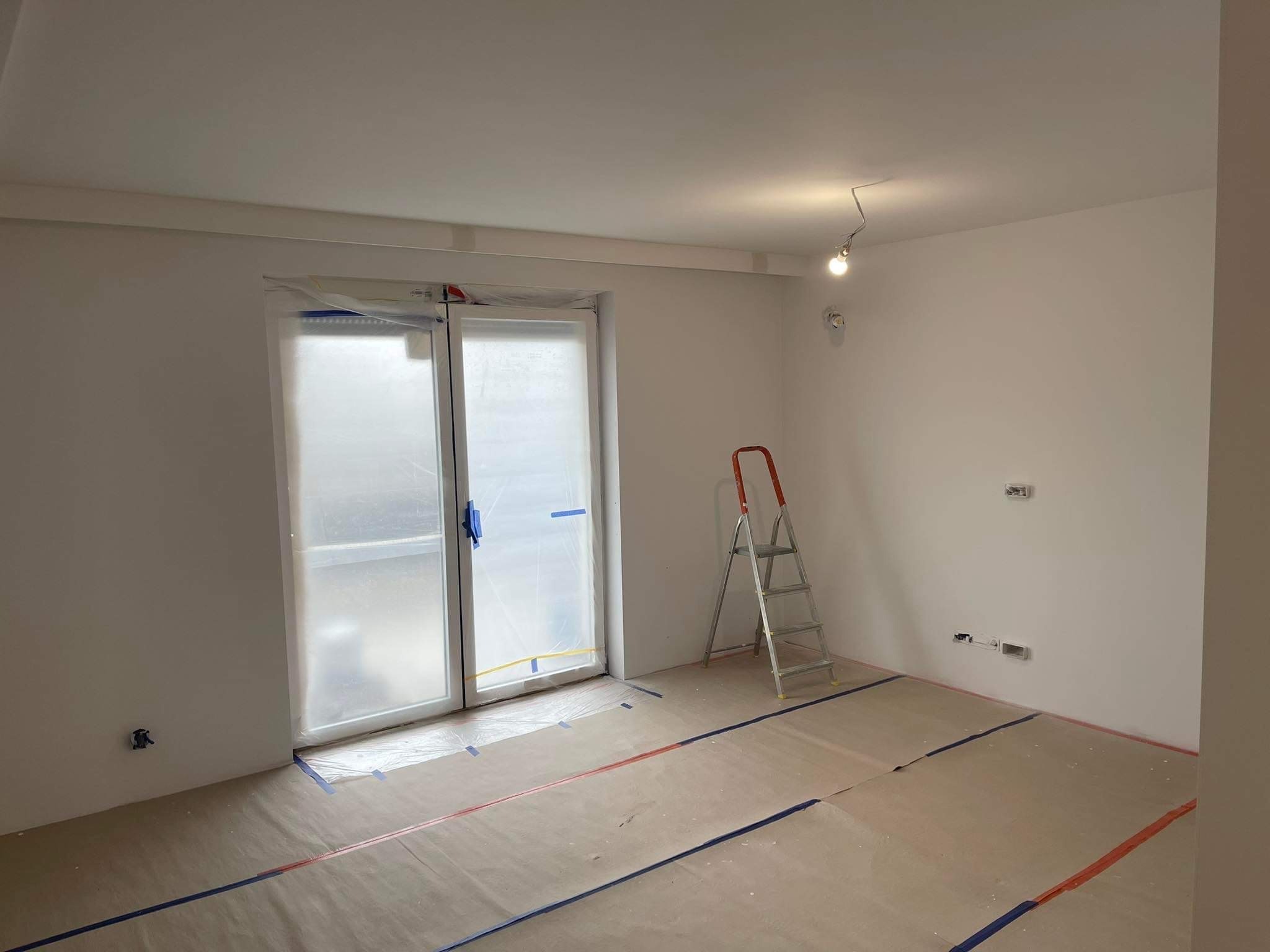 Pokój przygotowany do prac wykończeniowych: okno zabezpieczone folią, podłoga przykryta kartonem i taśmą malarską, drabina aluminiowa oparta o ścianę.