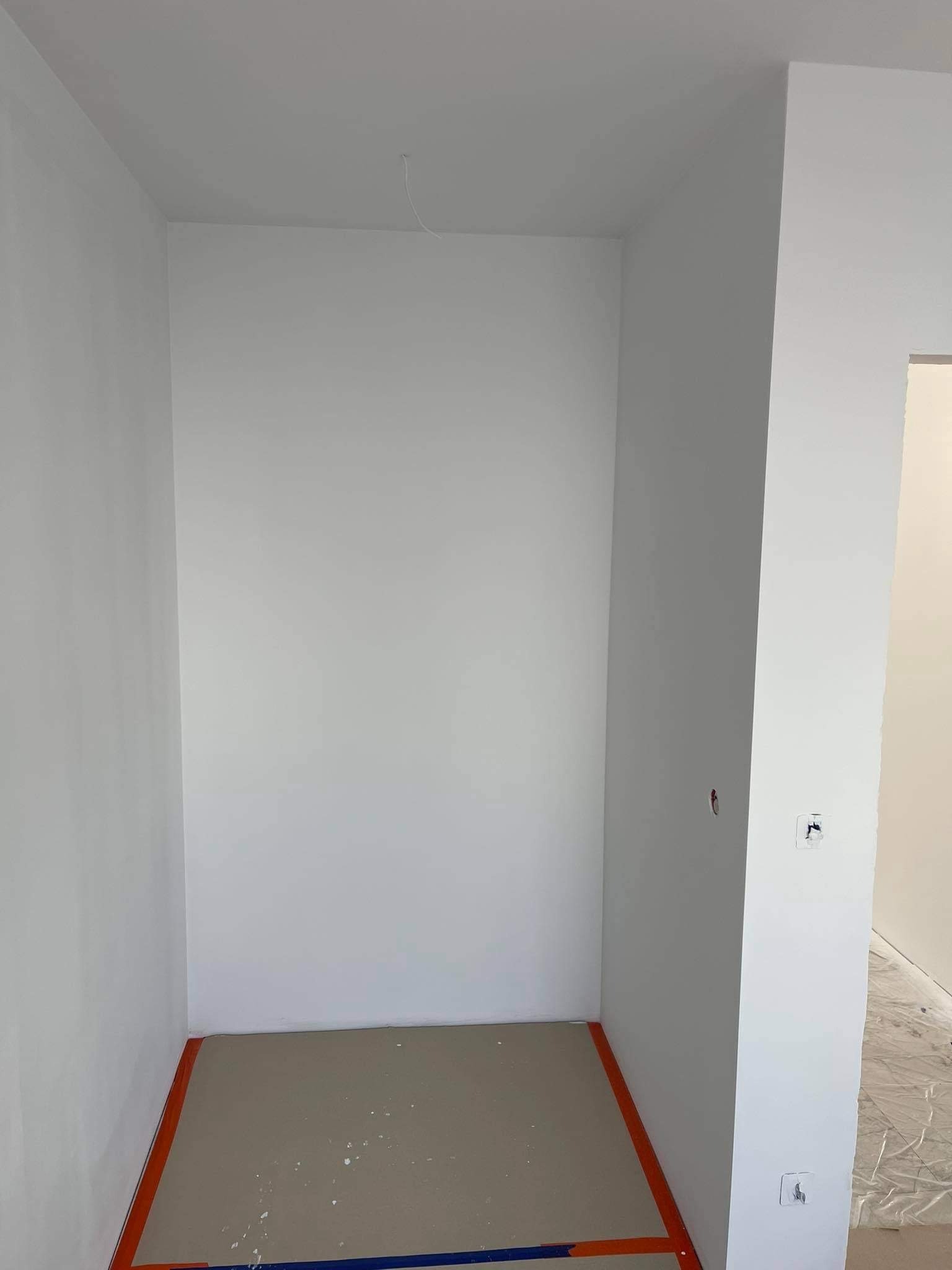 Pusta, świeżo pomalowana na biało przestrzeń z zabezpieczoną podłogą, widoczne ślady po pracach wykończeniowych i wystający kabel z sufitu.