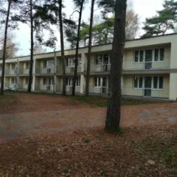 Dwukondygnacyjny budynek z balkonami i szarym pasem na elewacji, otoczony sosnami i brukowanym podjazdem pokrytym igliwiem.