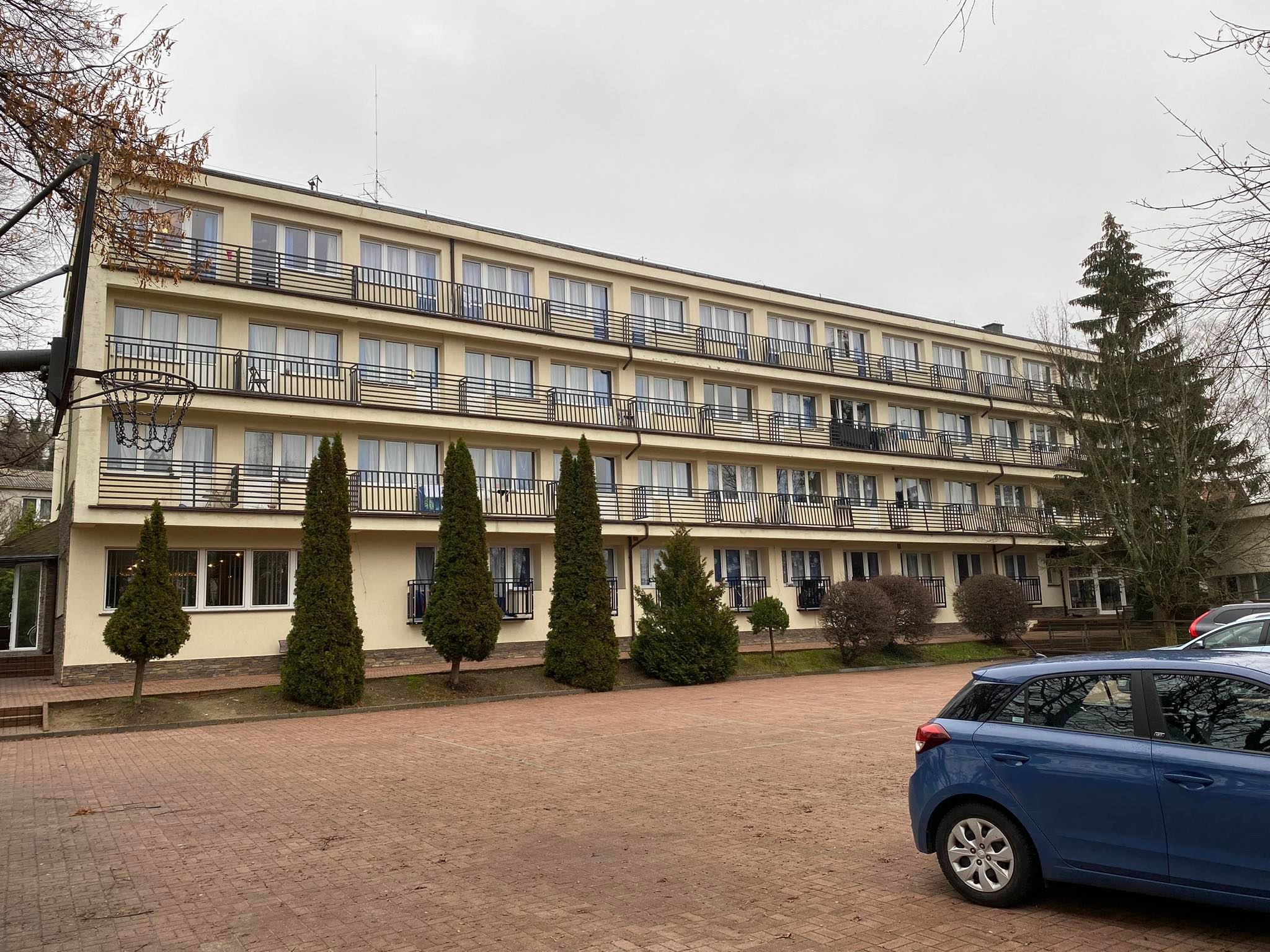 Czteropiętrowy budynek z balkonami, widok z parkingu z kostki brukowej, na pierwszym planie zaparkowany niebieski samochód.