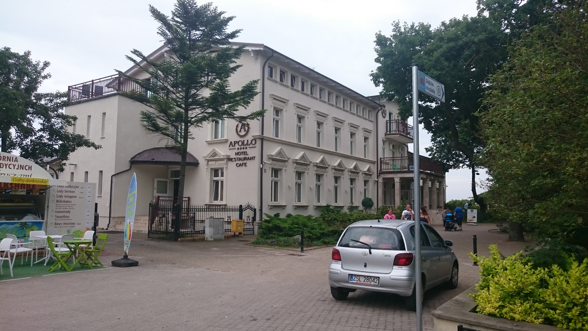 Elewacja czterogwiazdkowego hotelu Apollo z restauracją i kawiarnią, widoczna z ulicy z zaparkowanym srebrnym samochodem osobowym i straganem z lodami w tle.