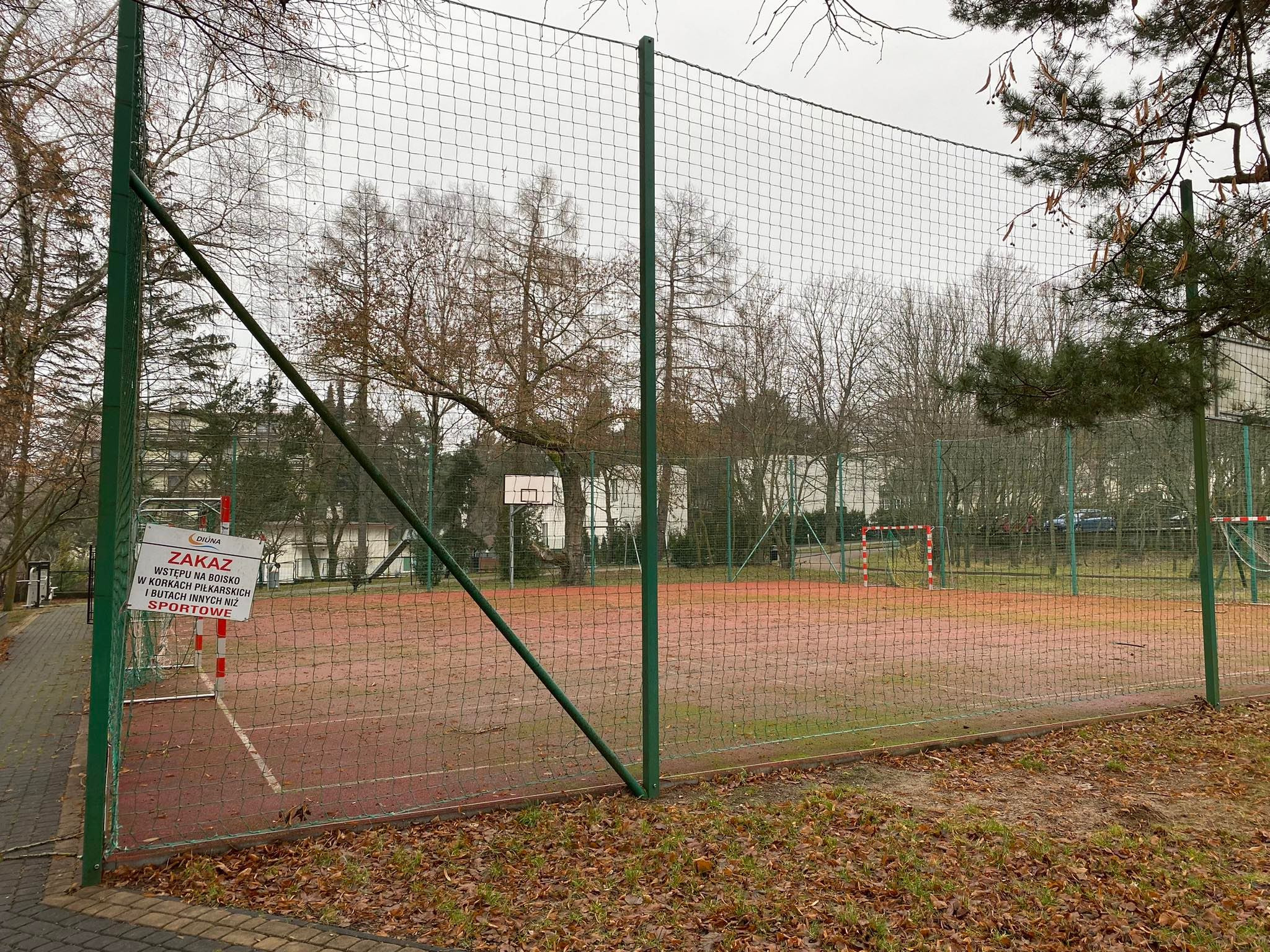 Ogrodzone boisko wielofunkcyjne z bramkami do piłki nożnej i koszem do koszykówki w tle, jesienne liście na ziemi, tablica z zakazem wstępu w obuwiu sportowym.