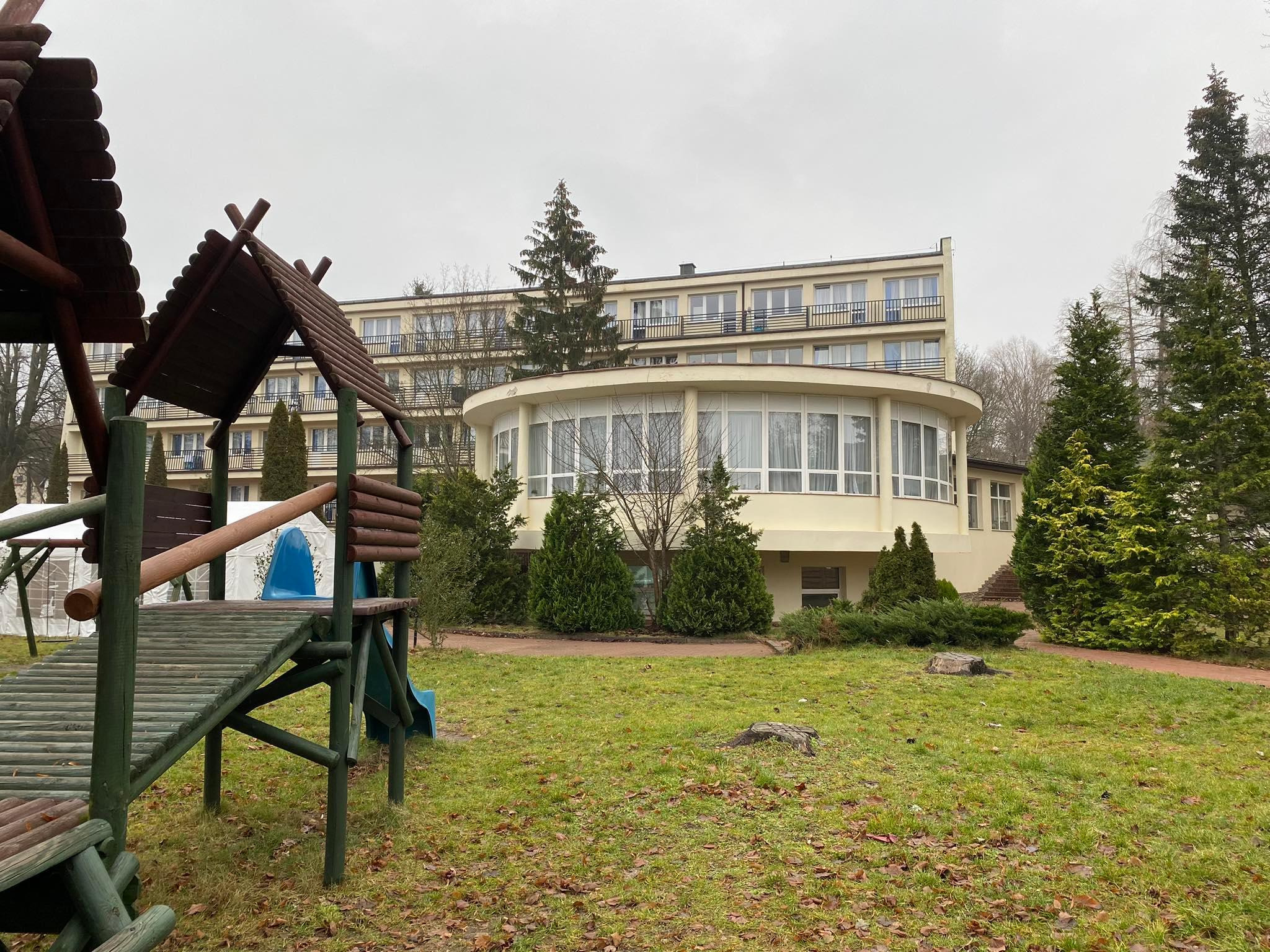 Żółty, trzypiętrowy budynek hotelowy z zaokrągloną częścią restauracyjną, widziany z perspektywy placu zabaw z drewnianą konstrukcją i zjeżdżalnią, na tle pochmurnego nieba.