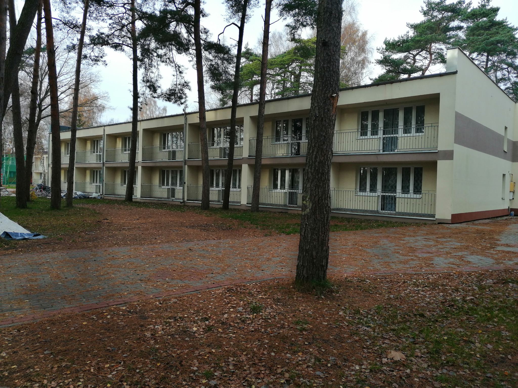 Dwukondygnacyjny budynek z balkonami i szarym pasem na elewacji, otoczony sosnami i brukowanym podjazdem pokrytym igliwiem.