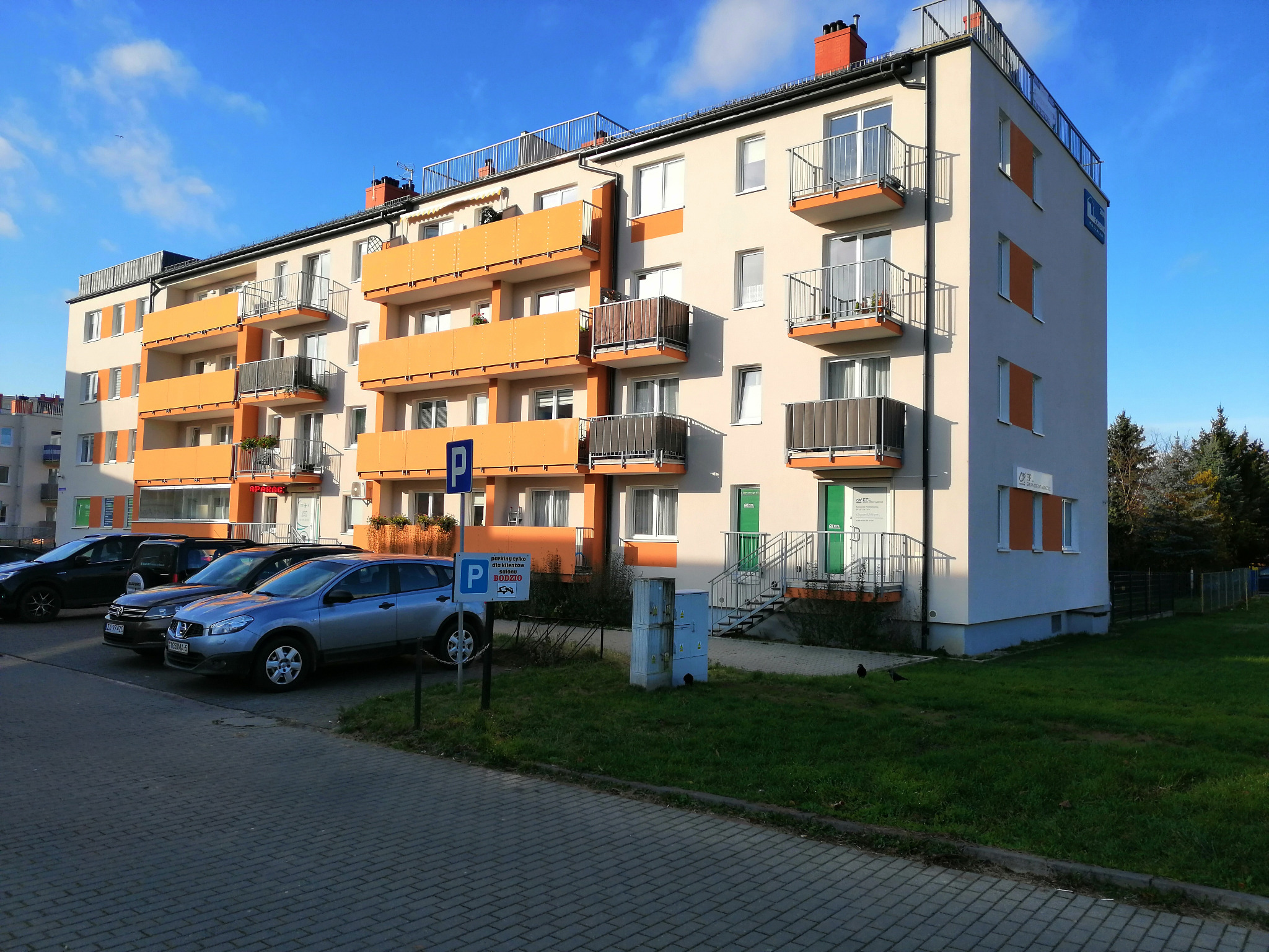 Nowoczesny budynek mieszkalny z pomarańczowymi balkonami i zaparkowanymi samochodami, widoczny znak parkingowy dla klientów salonu Bodzio.
