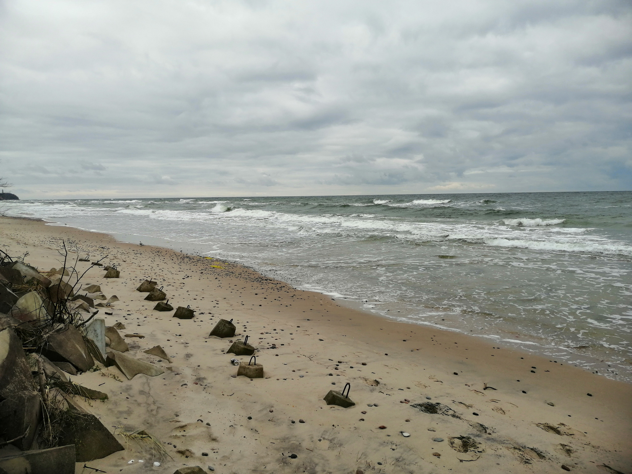 Plaża z betonowymi falochronami w kształcie sześcianów z metalowymi uchwytami, widok na morze i pochmurne niebo.