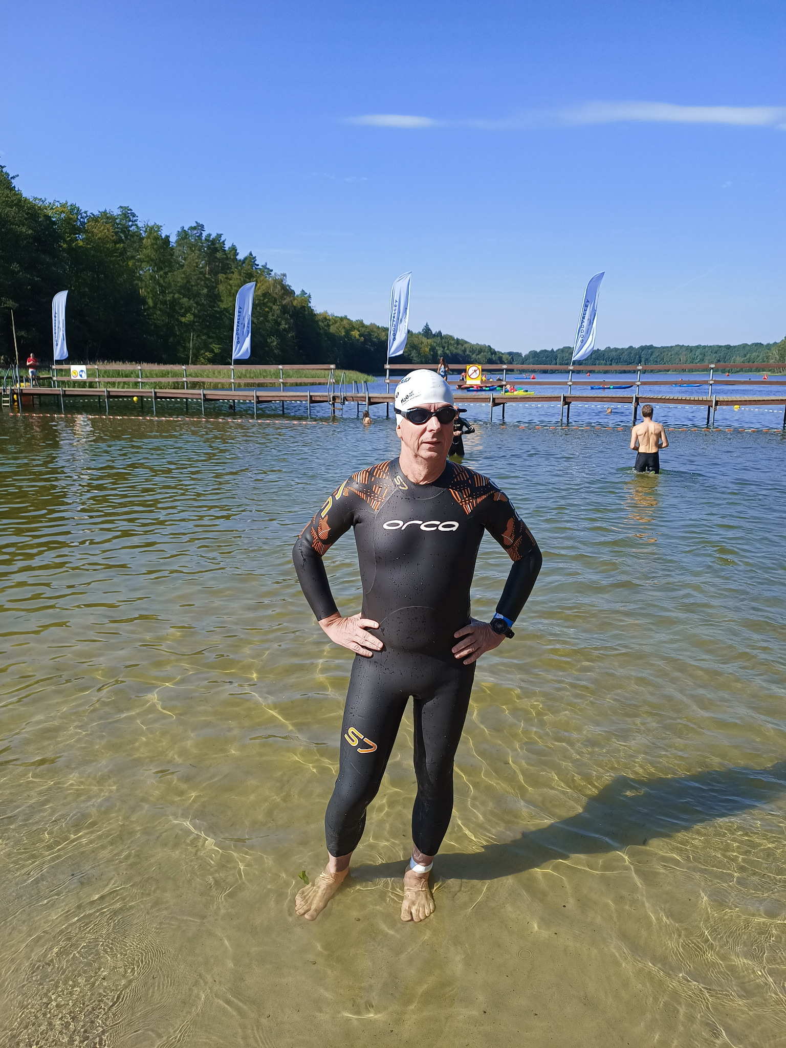 Mężczyzna w czarnym kombinezonie do triathlonu stoi w płytkiej, przejrzystej wodzie jeziora, z pomostem i flagami w tle, przygotowując się do zawodów.