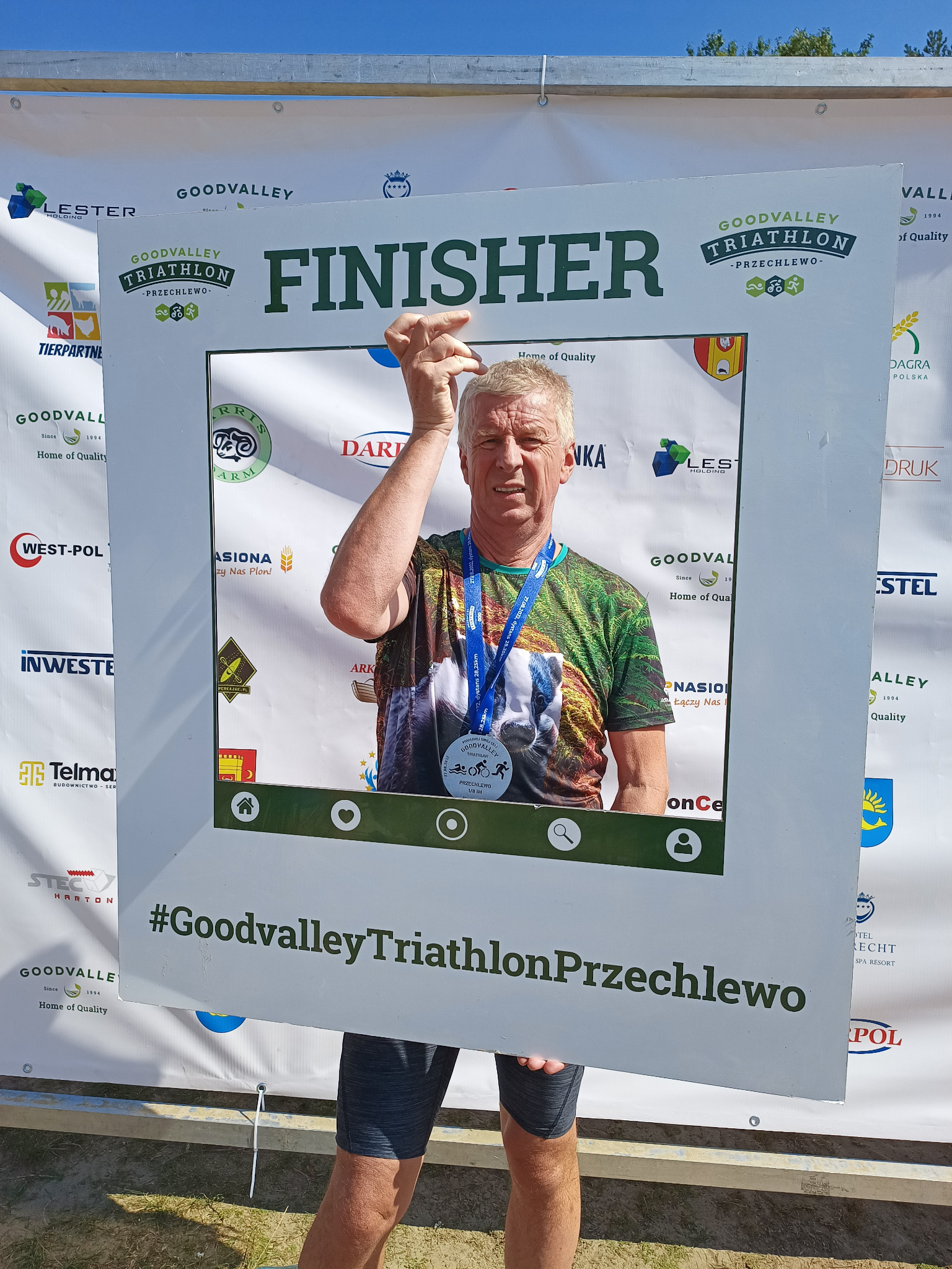 Mężczyzna z medalem na szyi pozuje w ramce z napisem FINISHER i logo Goodvalley Triathlon Przechlewo, na tle baneru sponsorskiego.