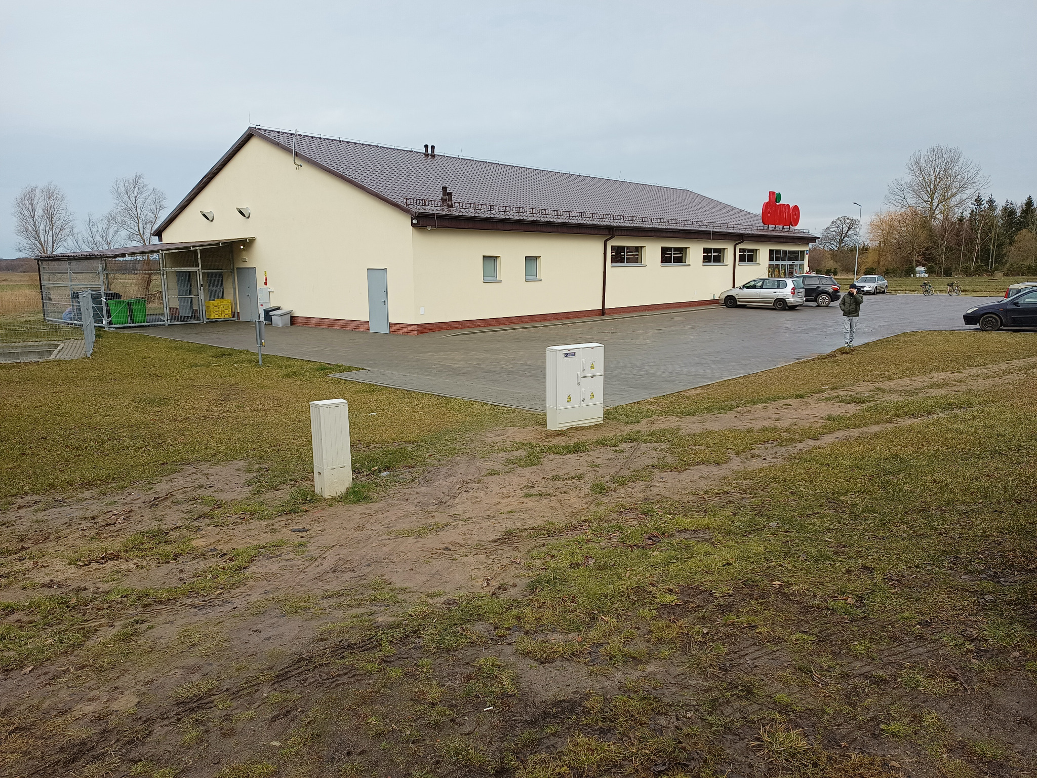 Parterowy budynek sklepu z brązowym dachem i czerwonym logo na elewacji, widoczny z zewnątrz na tle parkingu i drzew.