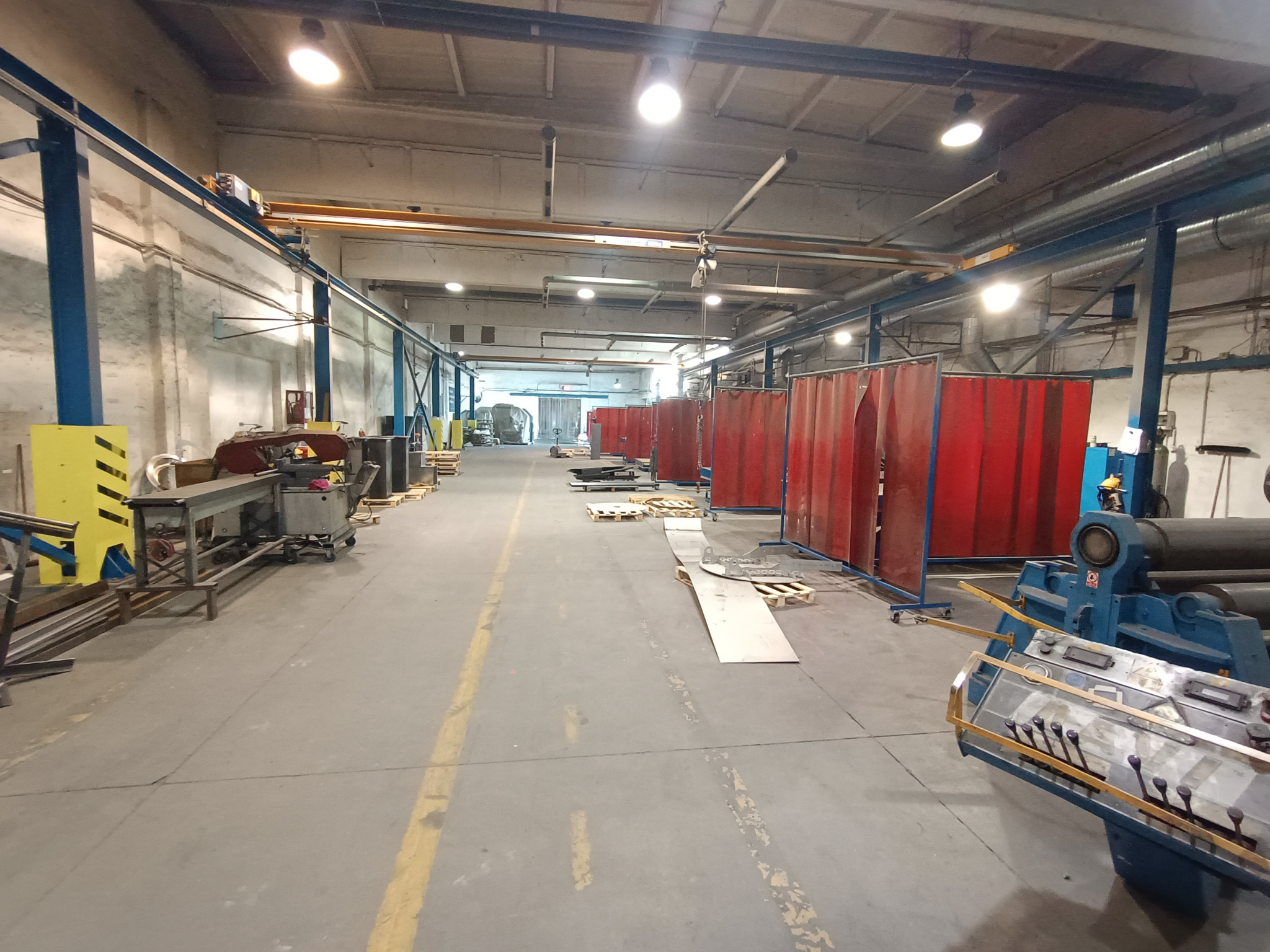 Wnętrze hali produkcyjnej z suwnicą, stołami warsztatowymi, parawanami spawalniczymi i maszyną do gięcia blach.