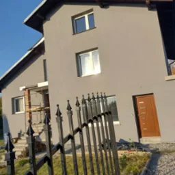 Dwupiętrowy dom z szarą elewacją w trakcie prac wykończeniowych, widoczne rusztowania przy wejściu, czarne metalowe ogrodzenie na pierwszym planie.