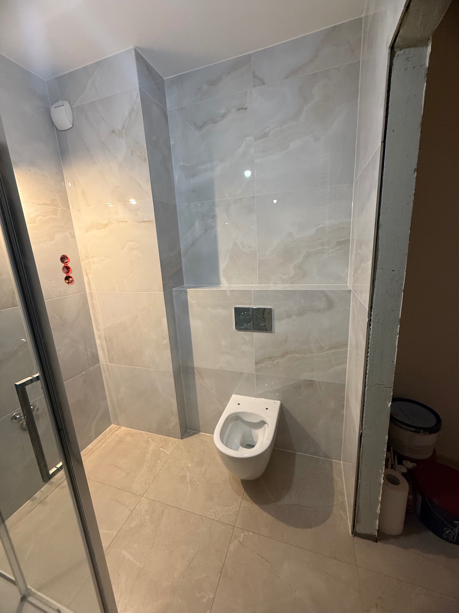 Wykończona łazienka z beżowymi płytkami imitującymi marmur, wisząca toaleta, widoczne elementy instalacji prysznicowej. Minimalistyczny design z akcentami chromu.