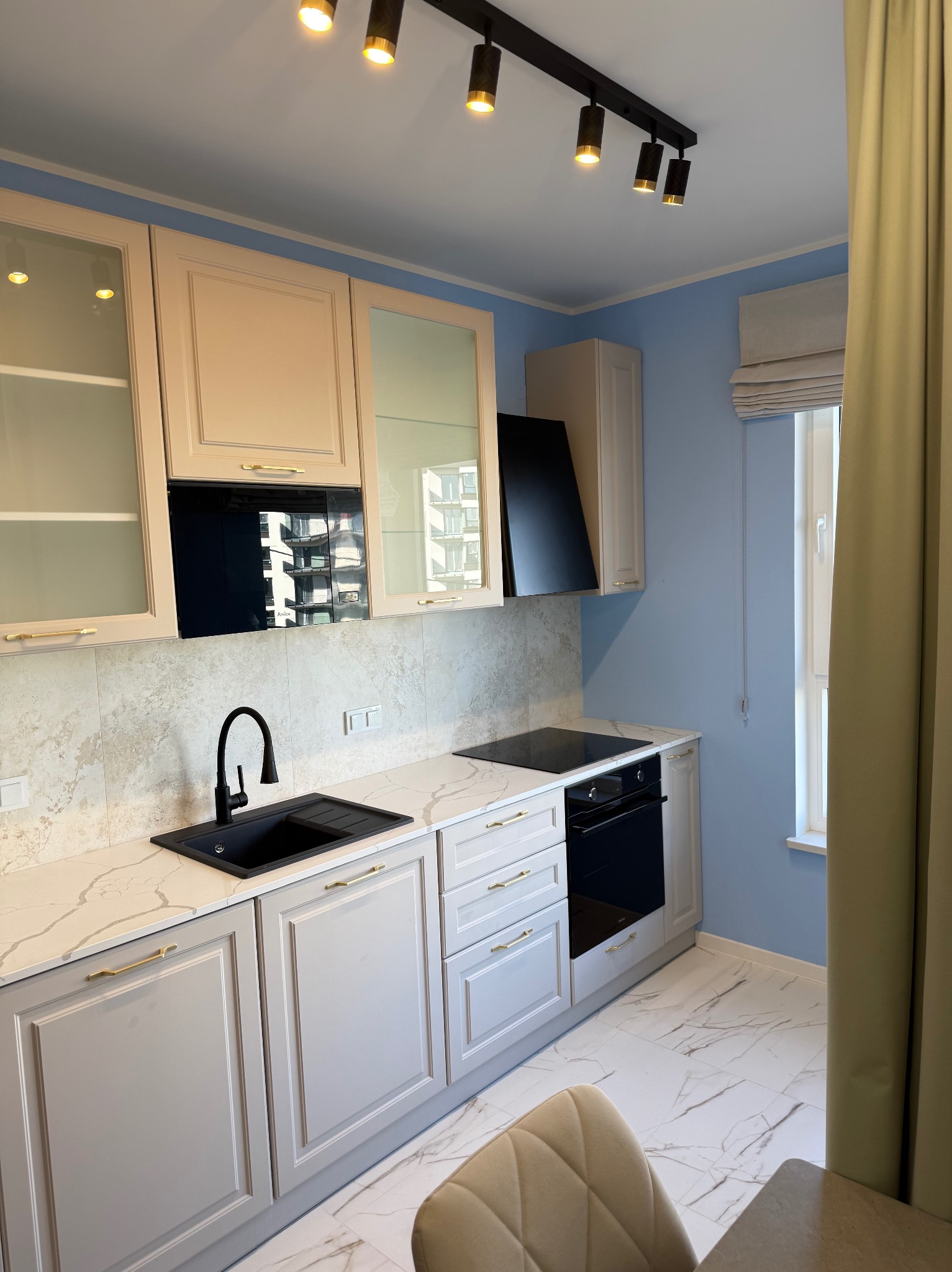 Elegancka kuchnia w odcieniach beżu i szarości z marmurowym blatem, czarną armaturą i oświetleniem punktowym. Błękitne ściany i zasłony dopełniają aranżację.