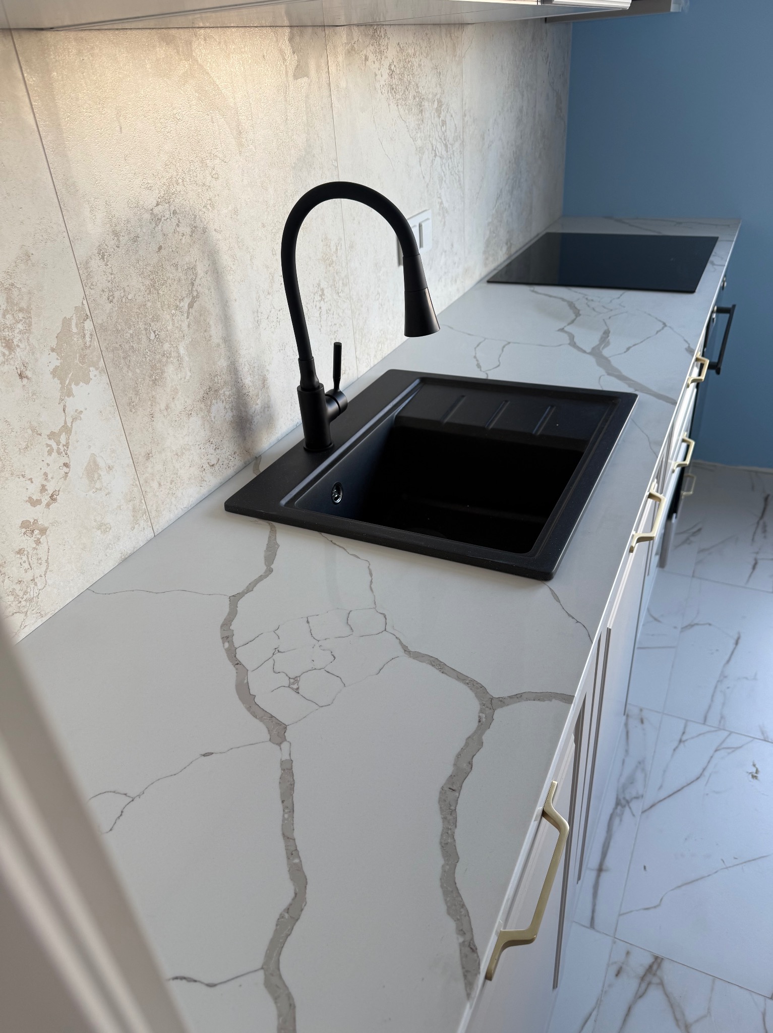 Nowoczesna kuchnia: czarny zlew i bateria na blacie z białego marmuru z szarymi żyłami, jasne płytki na ścianie i niebieska ściana w tle. Złote uchwyty szafek.