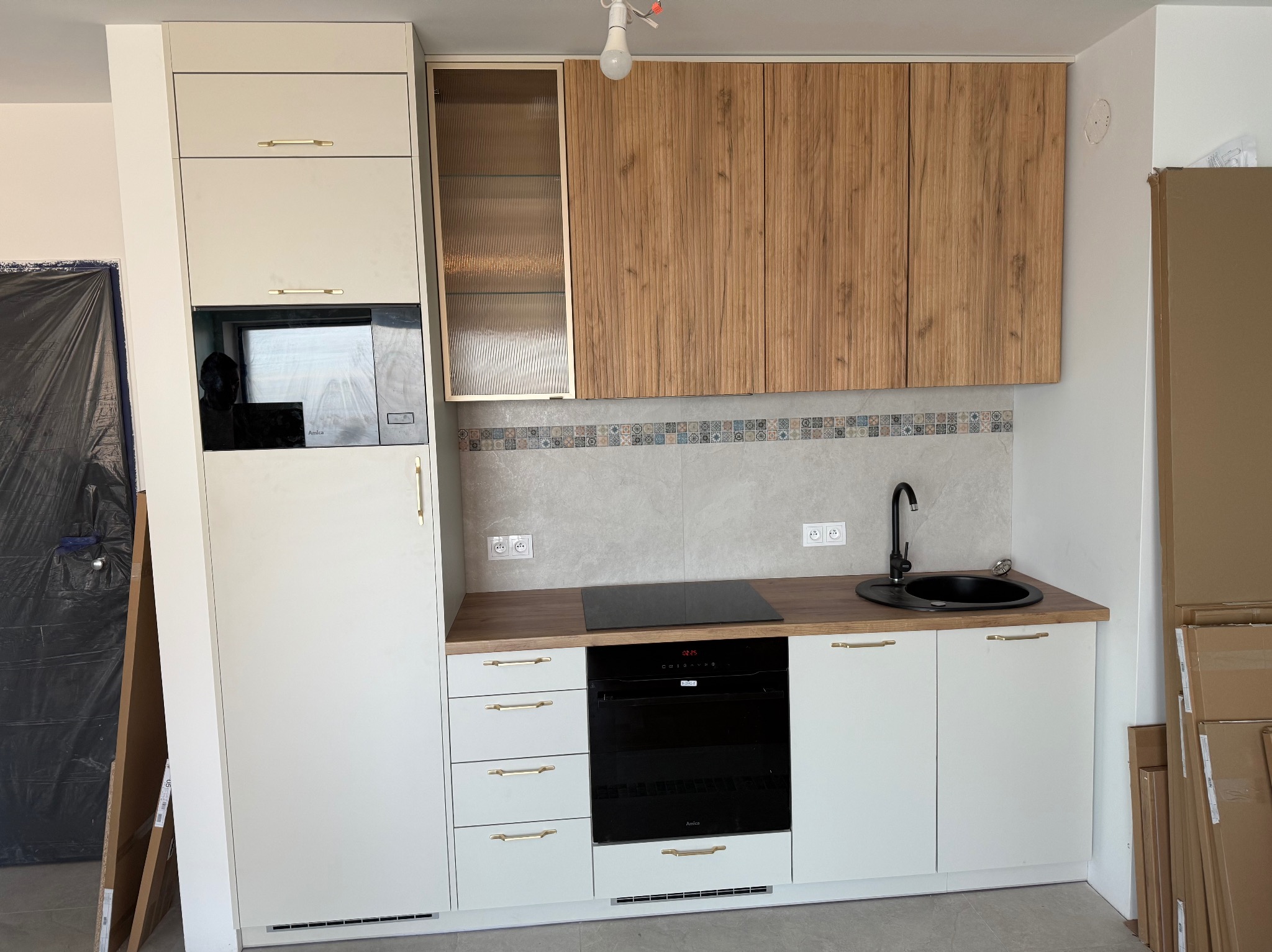 Nowoczesna kuchnia z białymi szafkami dolnymi i drewnopodobnymi górnymi, czarnym zlewem i płytą. Uwagę zwracają złote uchwyty i mozaika nad blatem.