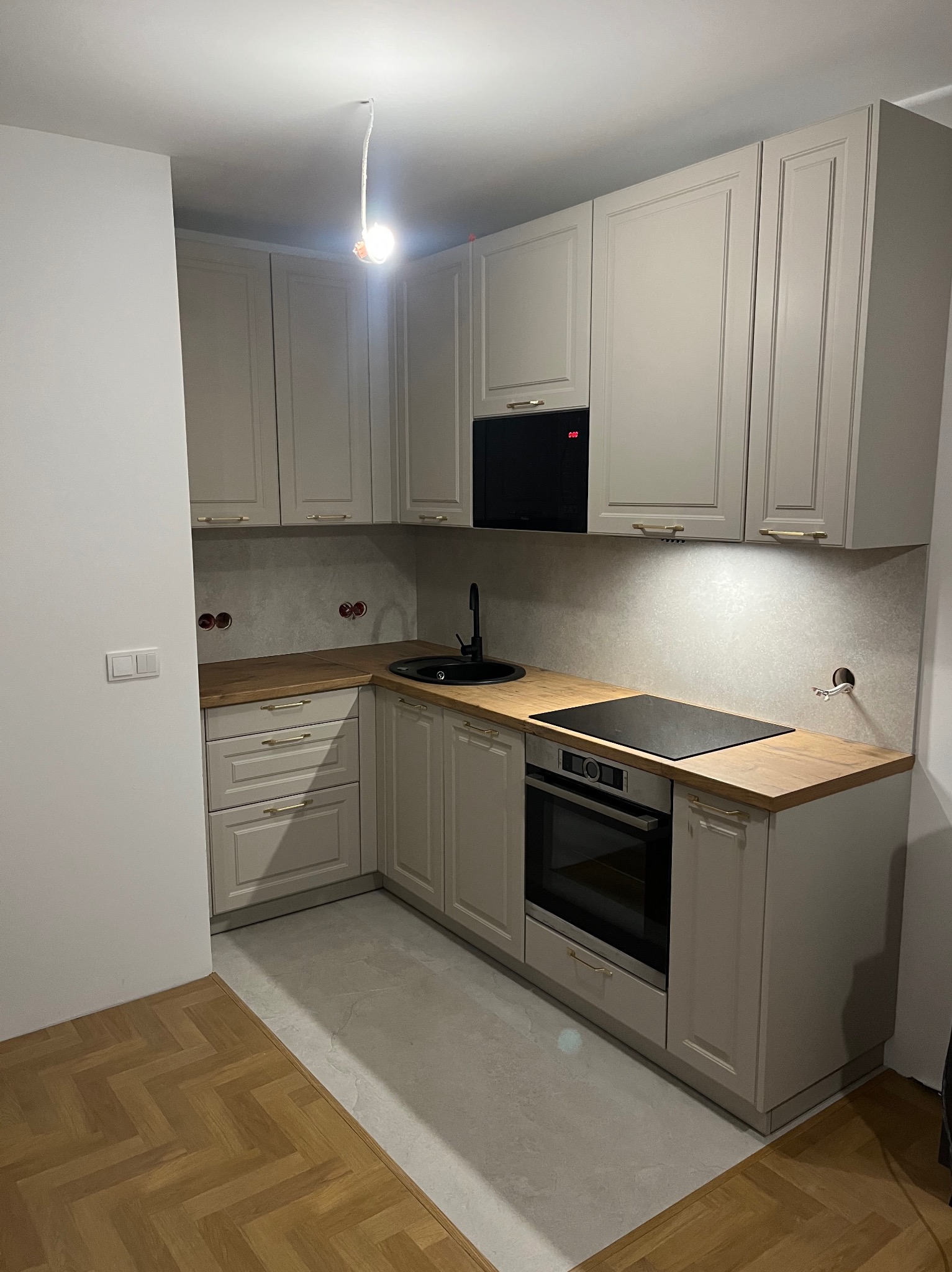 Jasna, narożna kuchnia z drewnianym blatem i czarnym zlewem. Widoczne gniazdka elektryczne i niedokończone oświetlenie sufitowe. Parkiet w jodełkę kontrastuje z szarą podłogą.