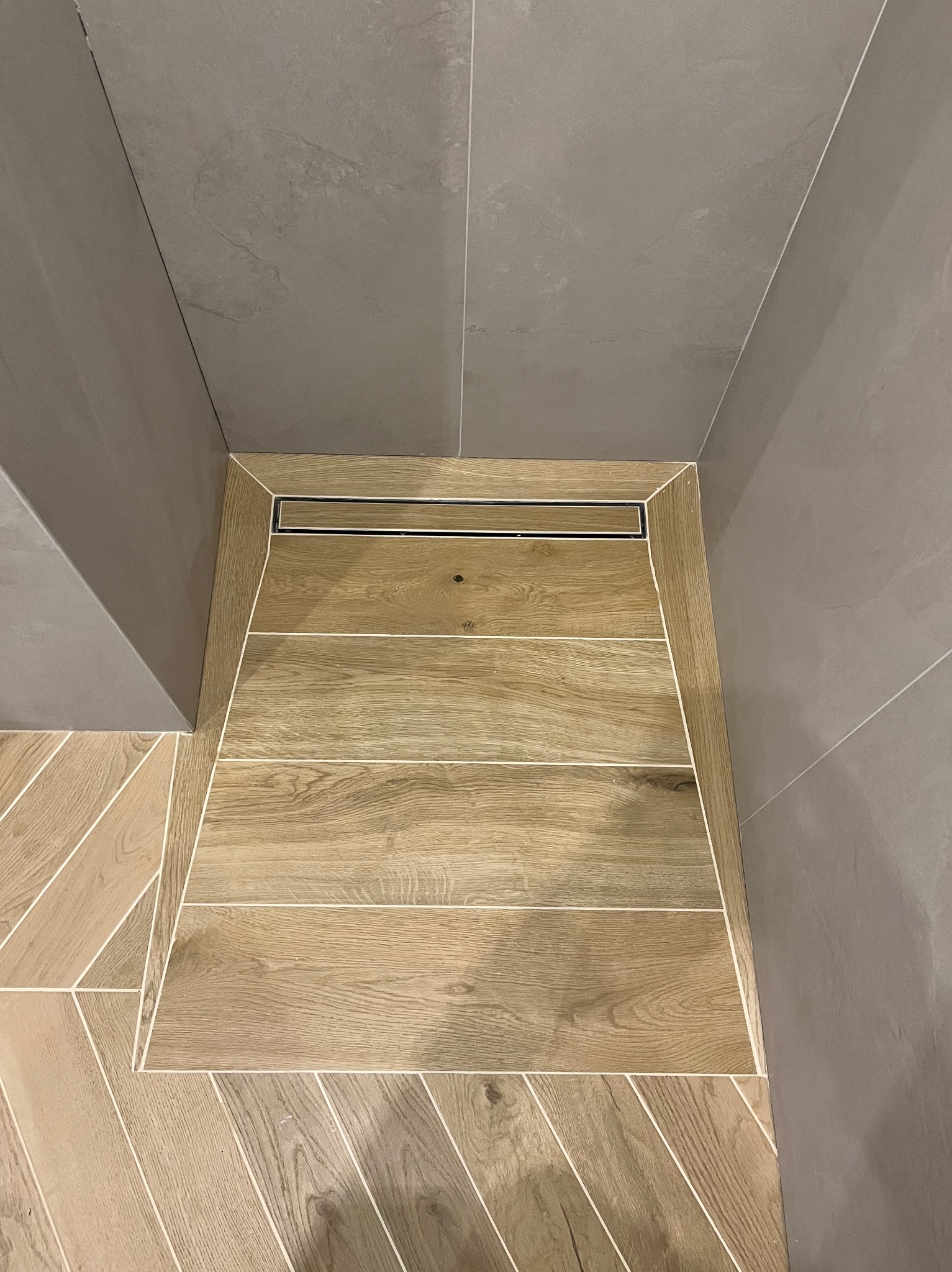 Nowoczesny odpływ liniowy w kabinie prysznicowej z drewnopodobnymi płytkami ułożonymi w jodełkę, szare płytki ścienne. Minimalistyczny design, precyzyjne wykończenie.