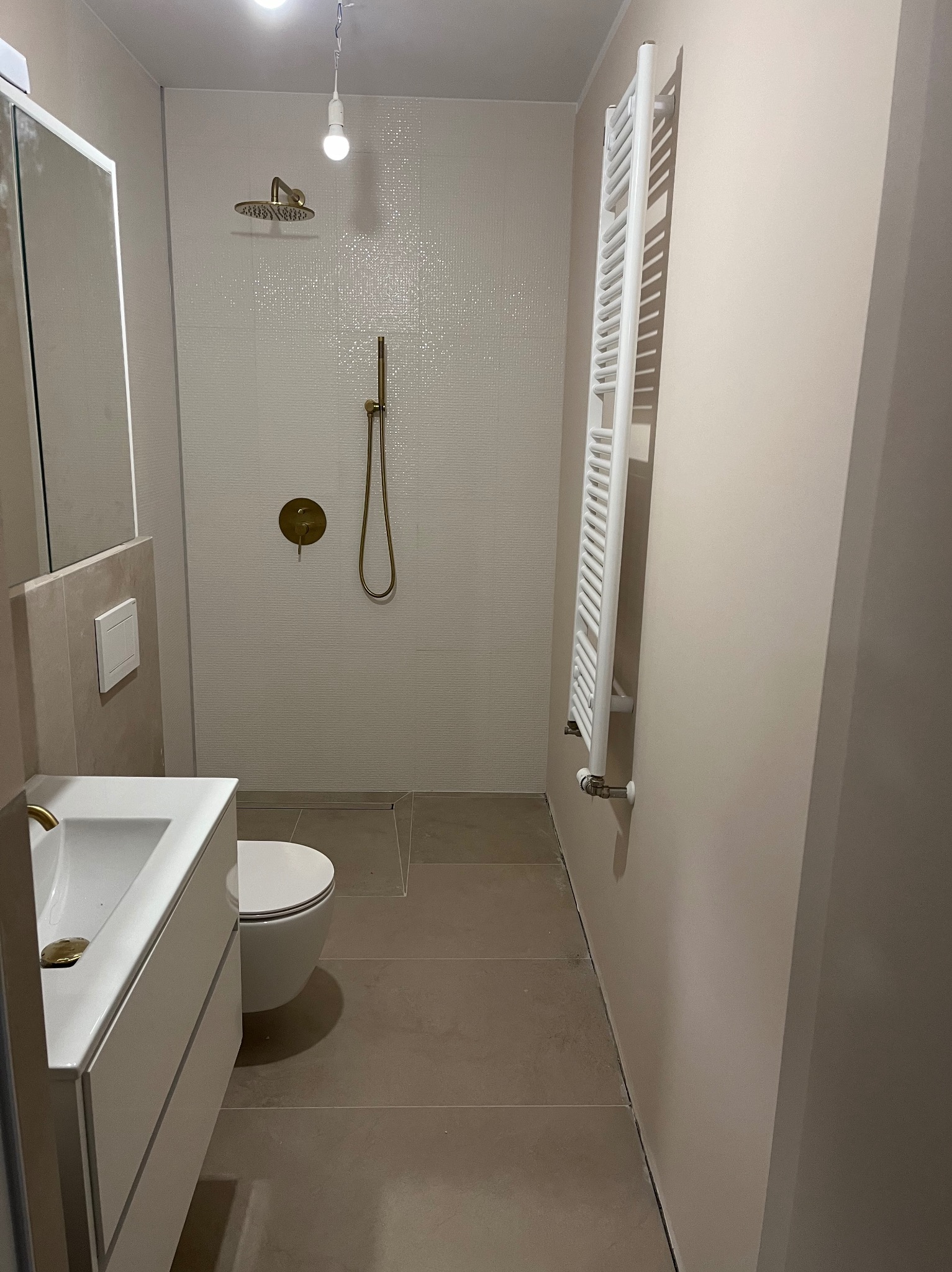 Wykończona łazienka z beżowymi płytkami, złotą armaturą i białym grzejnikiem drabinkowym. Umywalka z szafką, toaleta i kabina prysznicowa w minimalistycznym stylu.