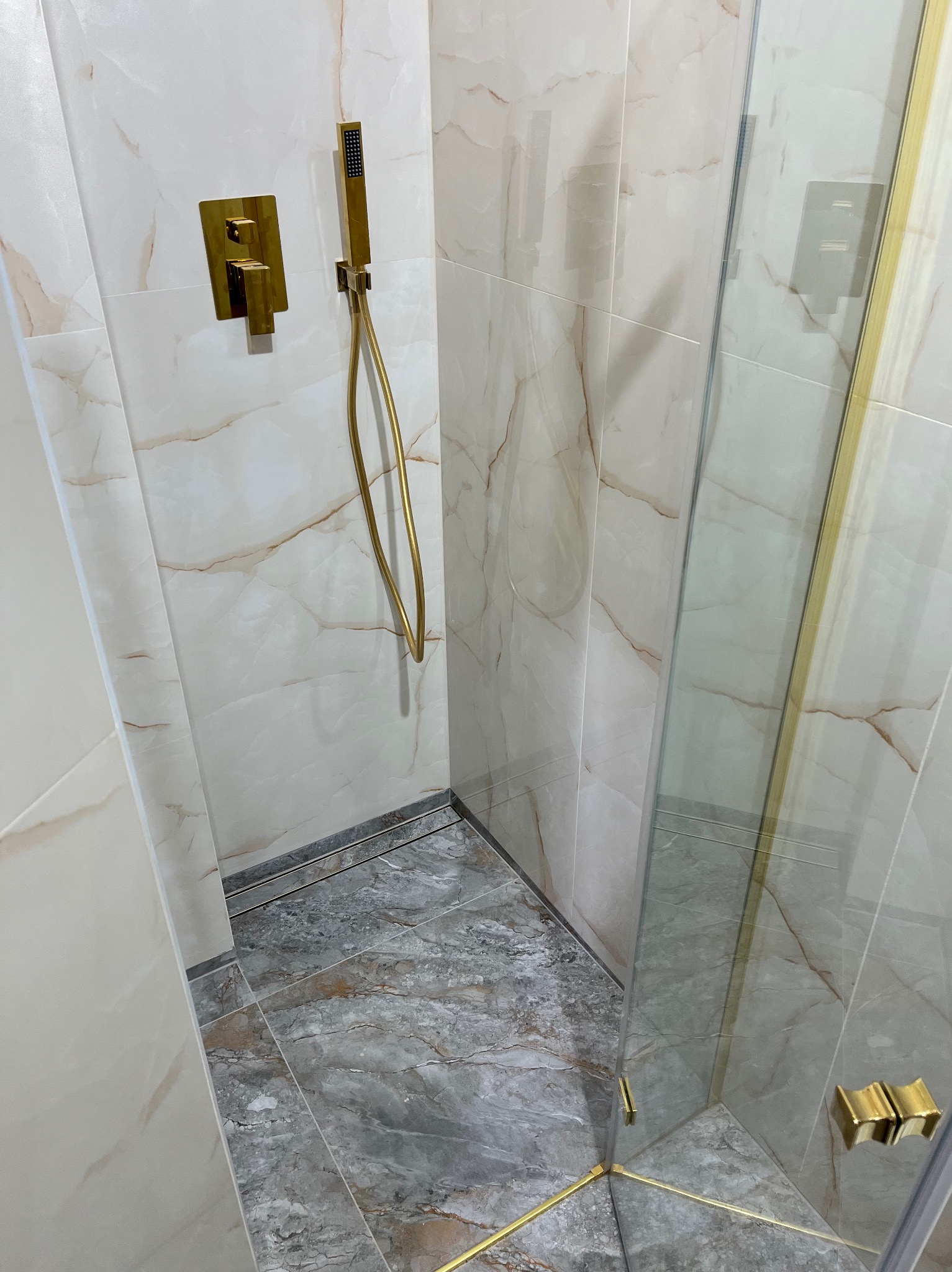 Nowoczesna kabina prysznicowa z marmurowymi płytkami i złotą armaturą. Szklane drzwi z eleganckimi, złotymi zawiasami. Minimalistyczny design.