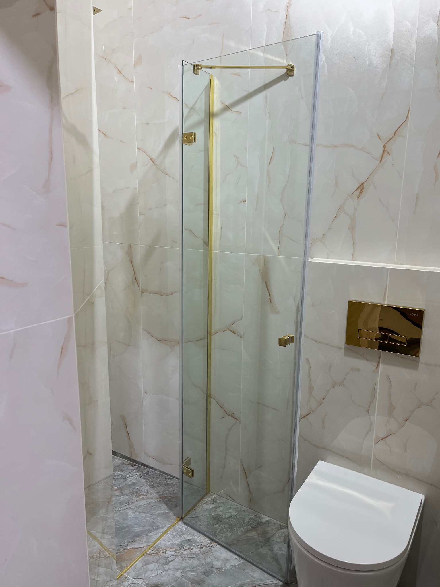 Wykończona łazienka z kabiną prysznicową ze złotymi detalami, marmurowe płytki na ścianach i podłodze, biała toaleta. Nowoczesny design.