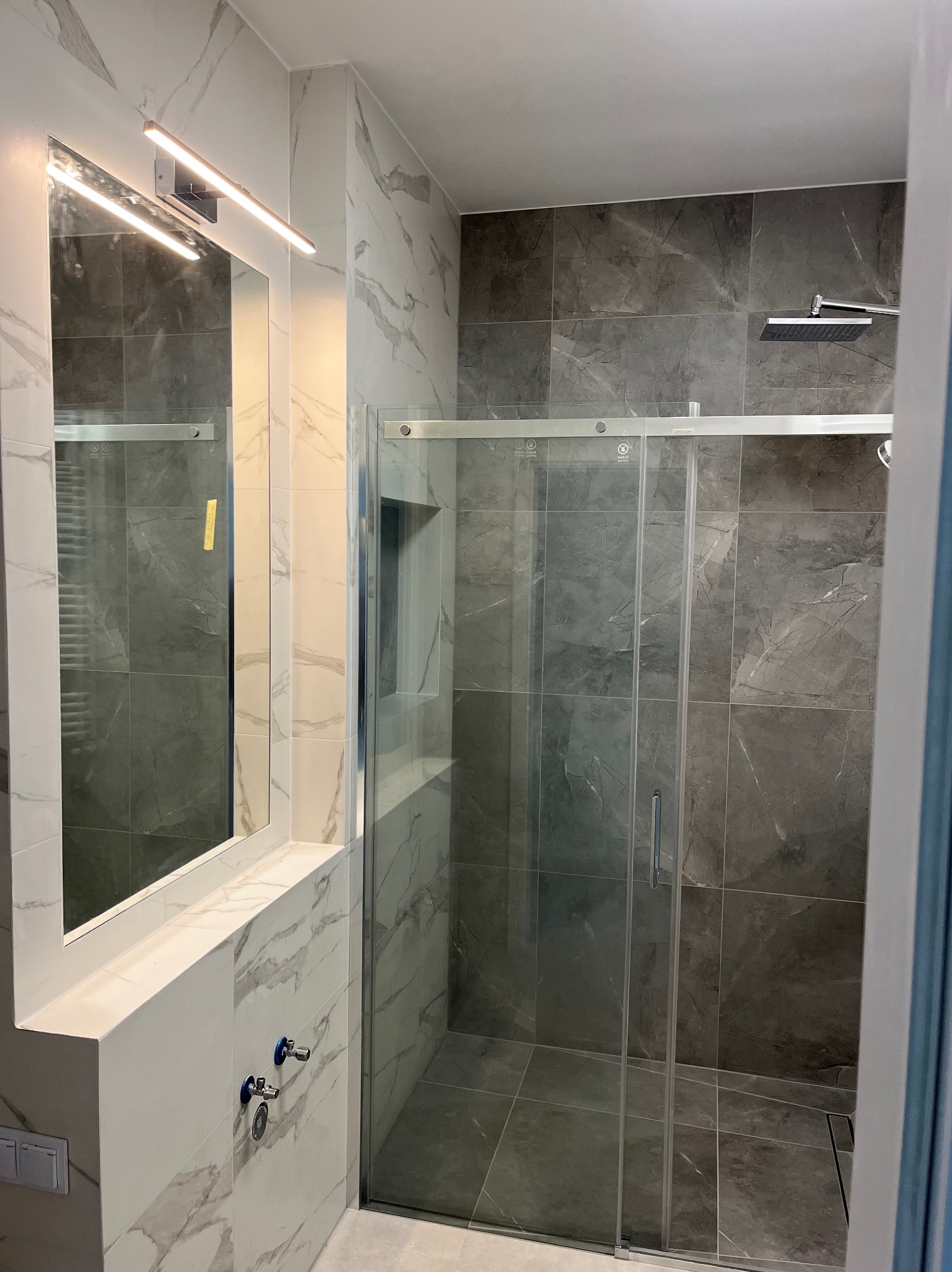 Nowoczesna łazienka z kabiną prysznicową typu walk-in, wykończona płytkami imitującymi marmur w odcieniach szarości i bieli. Widoczne chromowane elementy armatury i oświetlenie.