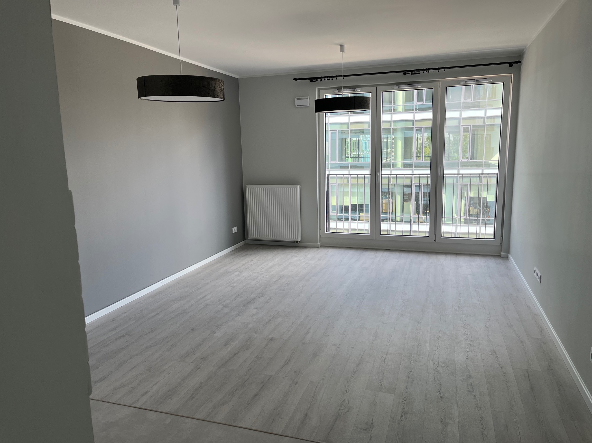 Pusty, szary pokój z panelami podłogowymi, grzejnikiem i dużymi oknami balkonowymi. Dwie lampy wiszące z czarnymi abażurami. Świeżo wykończone wnętrze.