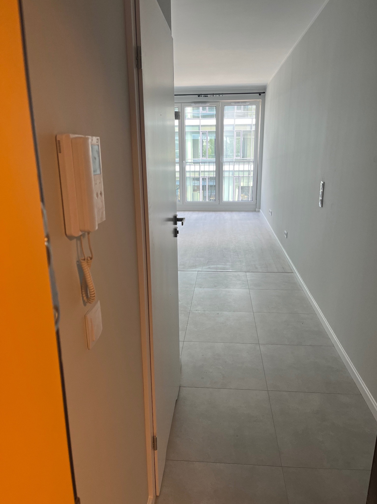 Jasne, wykończone wnętrze mieszkania z szarymi płytkami podłogowymi i białymi ścianami, widok zza uchylonych drzwi wejściowych z domofonem.