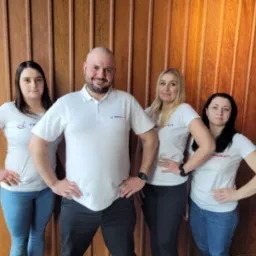 Grupa czterech osób (trzy kobiety i mężczyzna) w białych koszulkach z logo firmy 'Czysta Prawa 24' pozuje na tle ściany z drewnianych paneli w ciepłym oświetleniu.