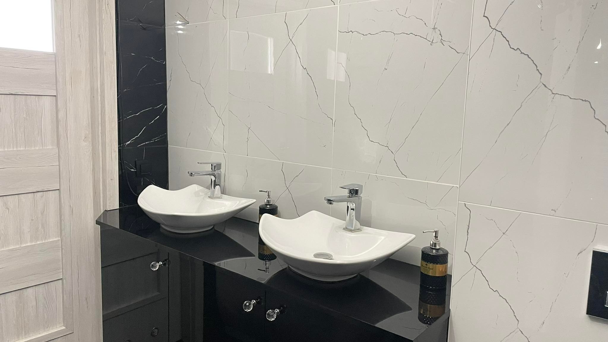 Nowoczesna łazienka z dwiema białymi umywalkami o falistym kształcie na czarnym, błyszczącym blacie z ozdobnymi uchwytami, wykończona białymi płytkami imitującymi marmur i czarnym pasem płytek...