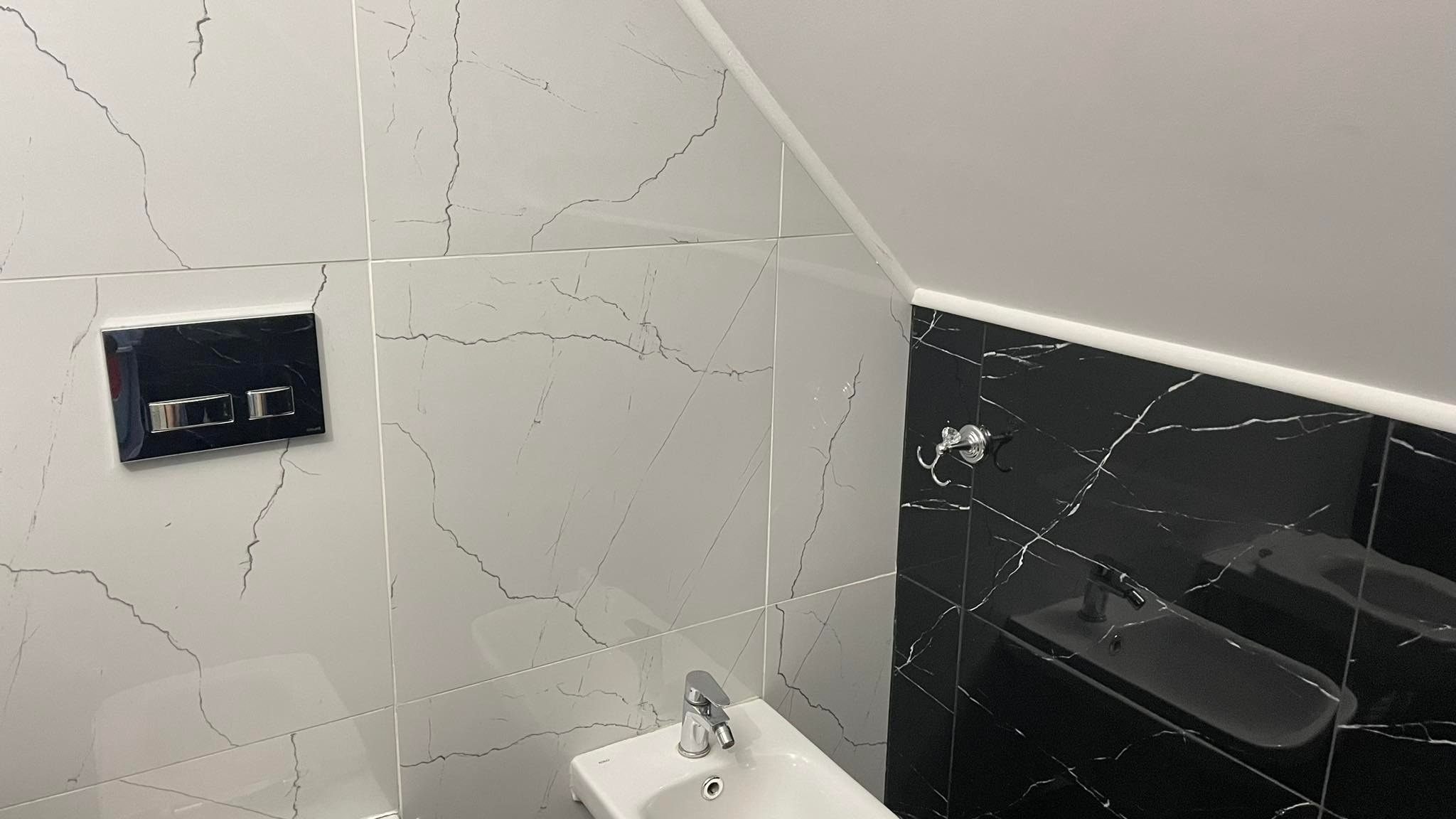 Biało-czarna łazienka z umywalką, chromowaną baterią i przyciskiem spłuczki w obudowie z czarnego szkła, wykończona płytkami imitującymi marmur