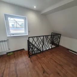 Nowoczesna balustrada metalowa z geometrycznym wzorem, zamontowana na antresoli z drewnianą podłogą, widok na okno i grzejnik.