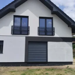 Nowoczesny dom z białymi ścianami, czarną dachówką i czarnymi, poziomymi balustradami balkonowymi z metalu. Okno na parterze z roletą zewnętrzną. Widoczne elementy wykończenia elewacji w kolorze...
