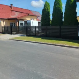 Nowoczesne, poziome ogrodzenie z metalowych profili w kolorze grafitowym, oddzielające posesję od ulicy, z widocznym fragmentem domu z czerwoną dachówką i białym samochodem dostawczym w tle.