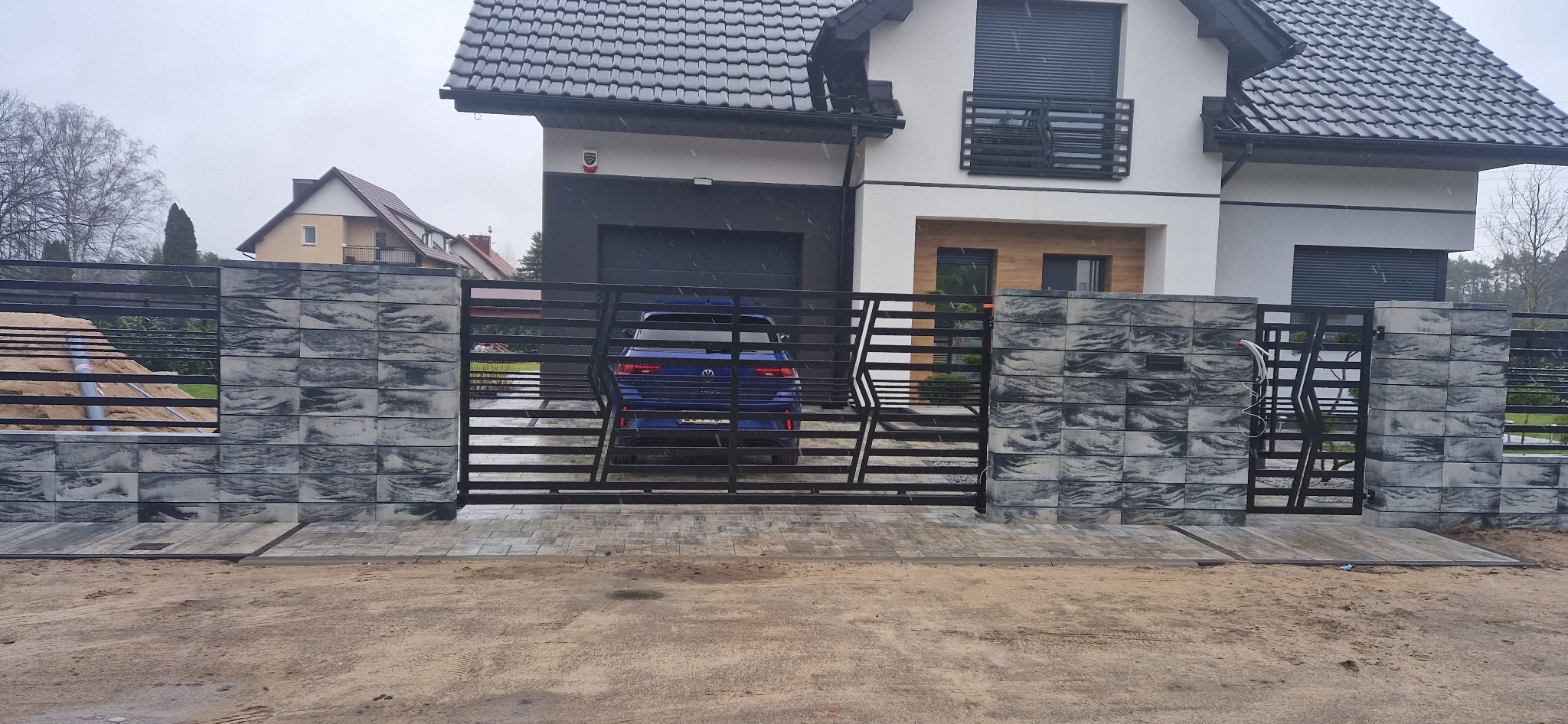 Nowoczesne ogrodzenie posesji z przesuwną bramą metalową i kamiennymi słupkami w odcieniach szarości, na tle domu z ciemną dachówką i białymi ścianami, widok z zewnątrz na podjazd.