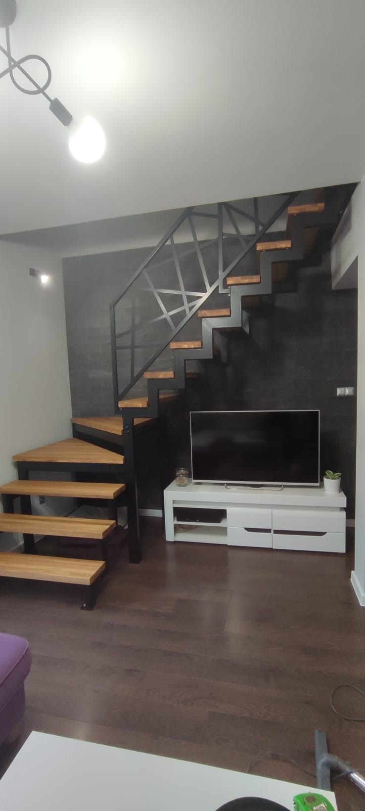 Nowoczesne schody z drewnianymi stopniami i czarną, geometryczną balustradą z metalu, umieszczone w przestronnym wnętrzu z telewizorem na białej szafce.
