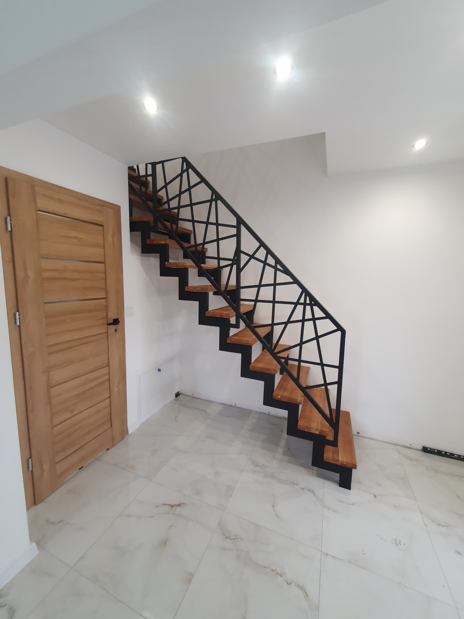Nowoczesne schody z drewnianymi stopniami i czarną, geometryczną balustradą w minimalistycznym wnętrzu z marmurową podłogą i drewnianymi drzwiami.